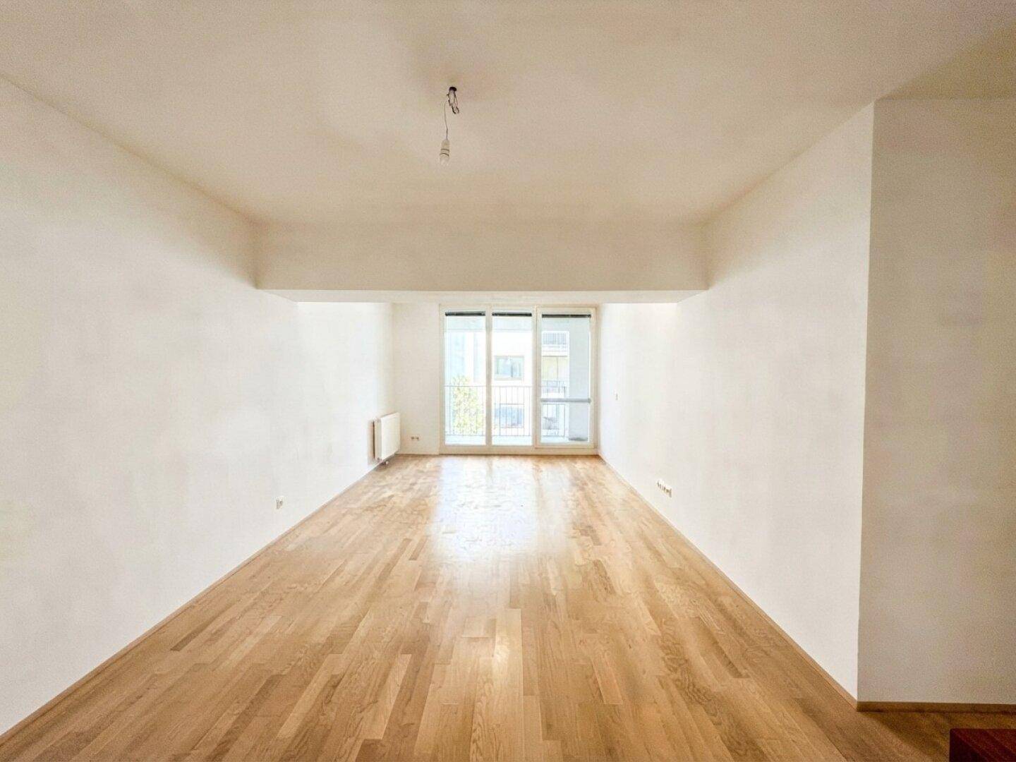 Helles, leeres Wohnzimmer mit Holzboden und Zugang zu einem Balkon durch große Fenster.