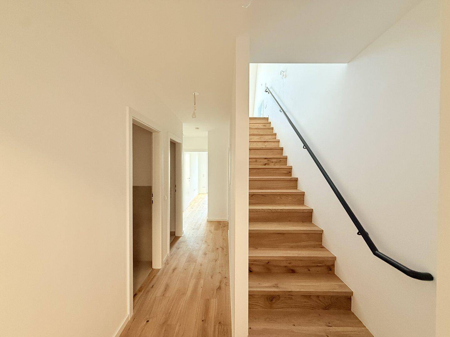 Heller Flur mit Holztreppe und schwarzem Geländer, führt zu den oberen Etagen.