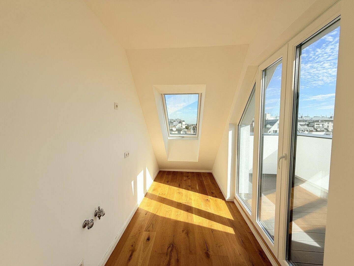 Schmalerer Bereich des Dachgeschosszimmers mit Holzboden, großem Fenster und Zugang zum Balkon.
