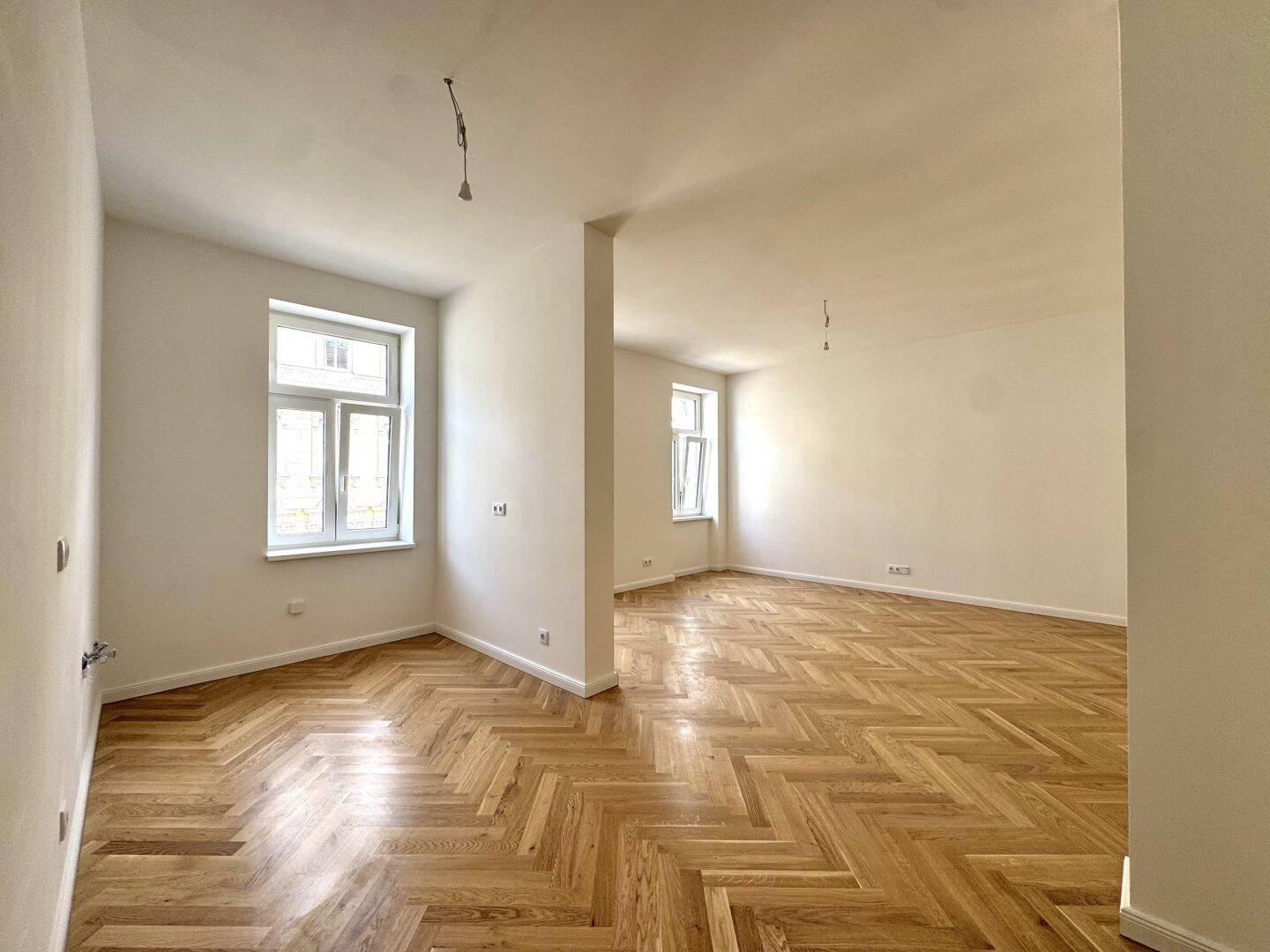 Geräumiges, lichtdurchflutetes Zimmer mit klassischem Fischgrätparkett und zwei großen Fenstern.