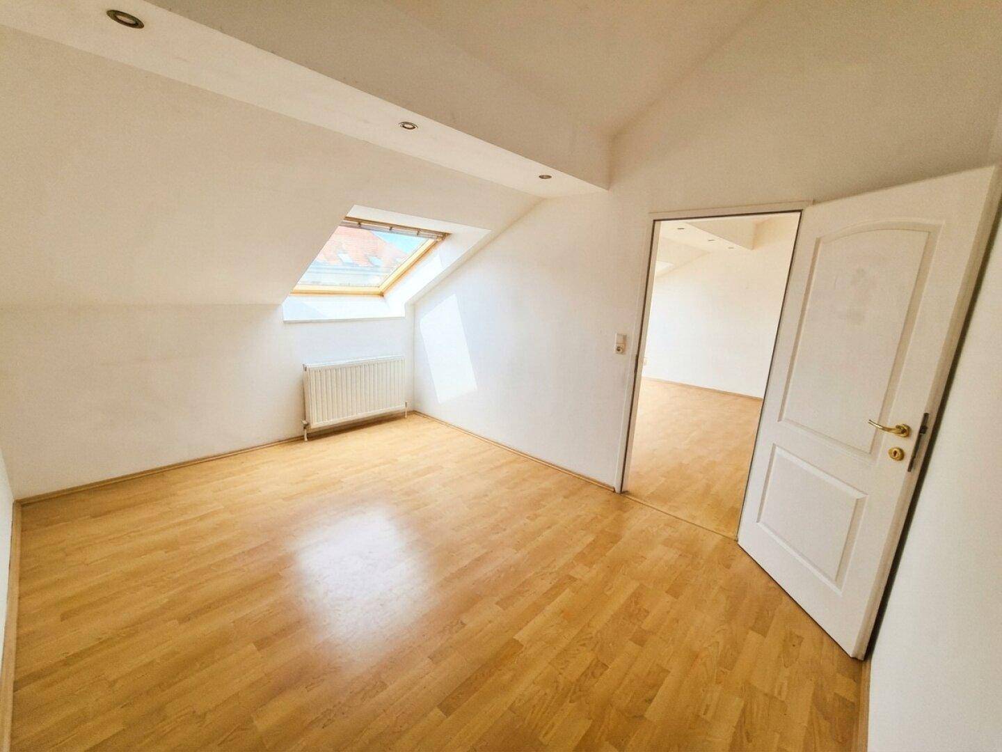 Geräumiges Zimmer im Dachgeschoss mit Holzfußboden, Dachfenster und Zugang zu einem weiteren Raum.