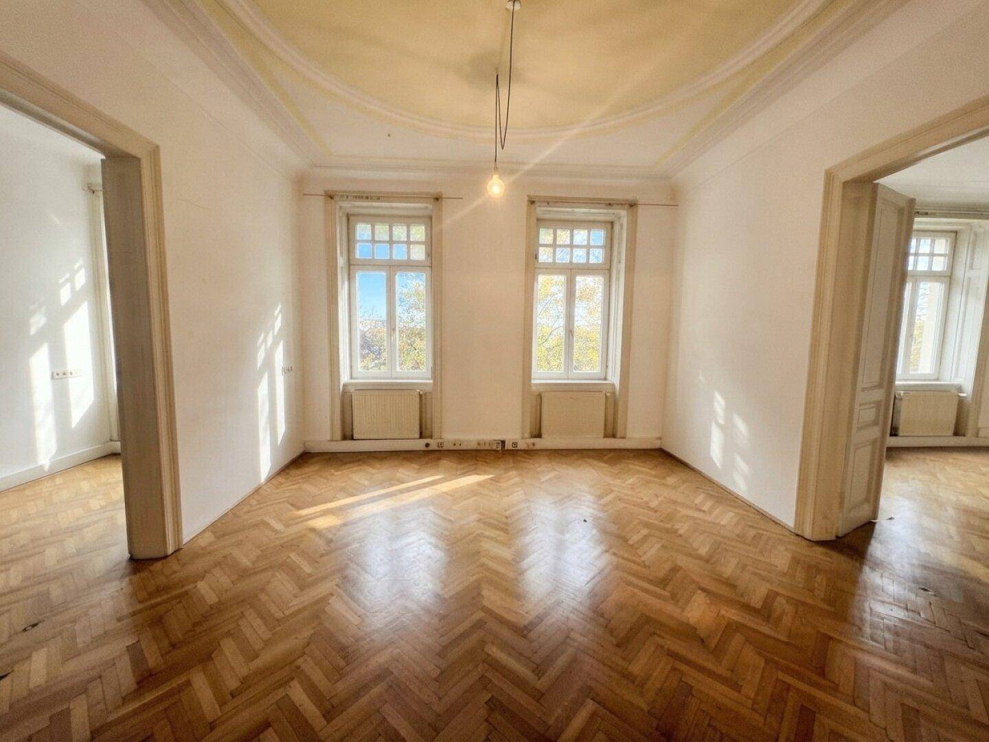 Lichtdurchfluteter Raum mit elegantem Fischgrätparkett und zwei hohen Fenstern, bereit zur Gestaltung.