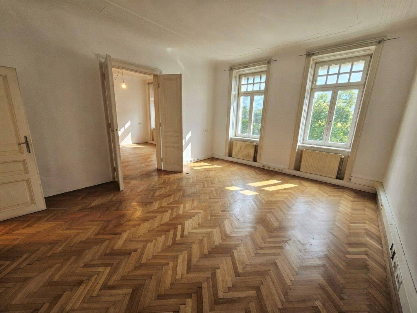 Geräumiges Zimmer mit sonnigem Fischgrätparkett, hohen Decken und Blick ins Grüne durch die Fenster.