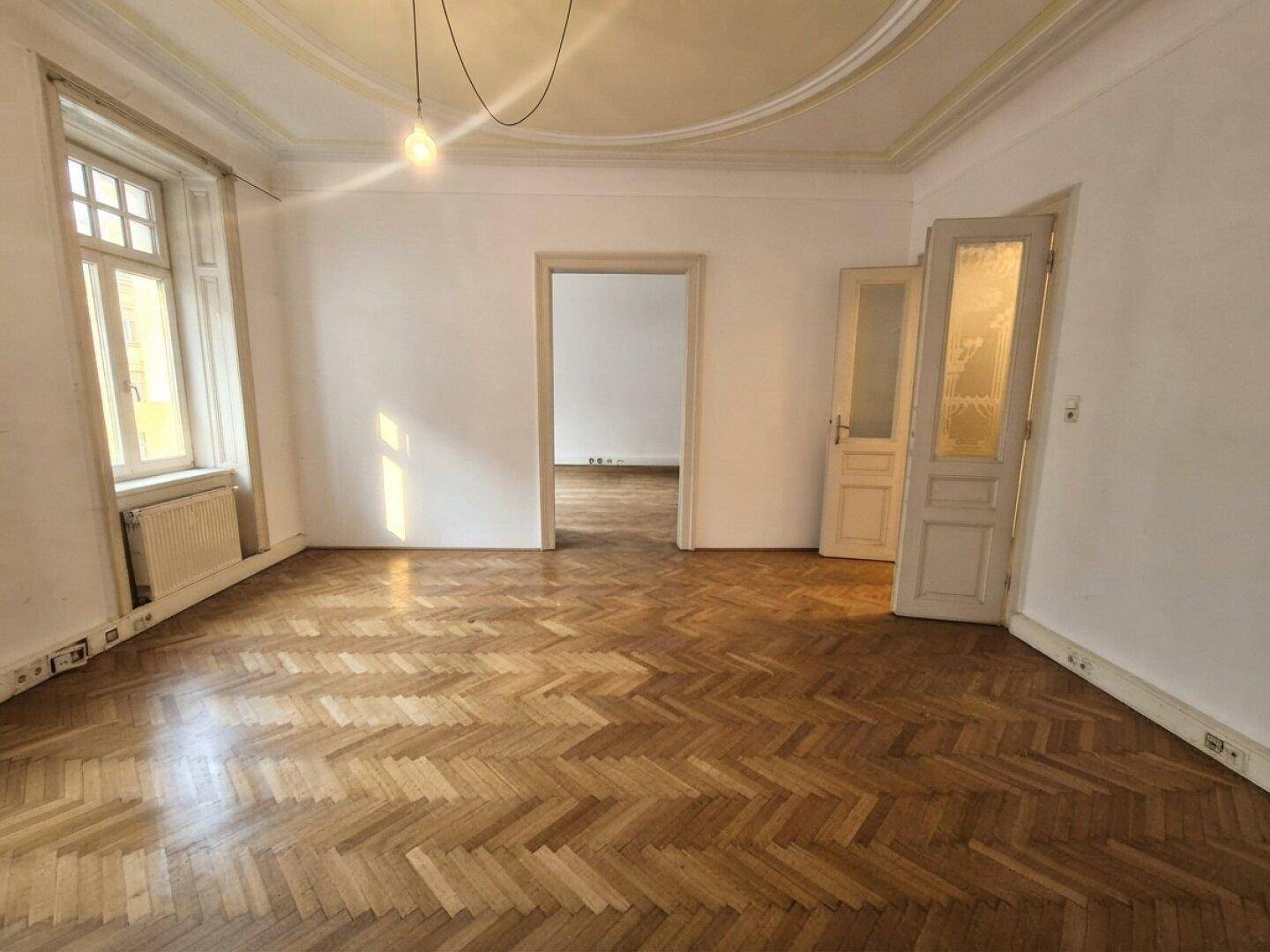 Helles, leeres Zimmer mit klassischem Fischgrätparkett, hohen Decken und Stuckelementen.