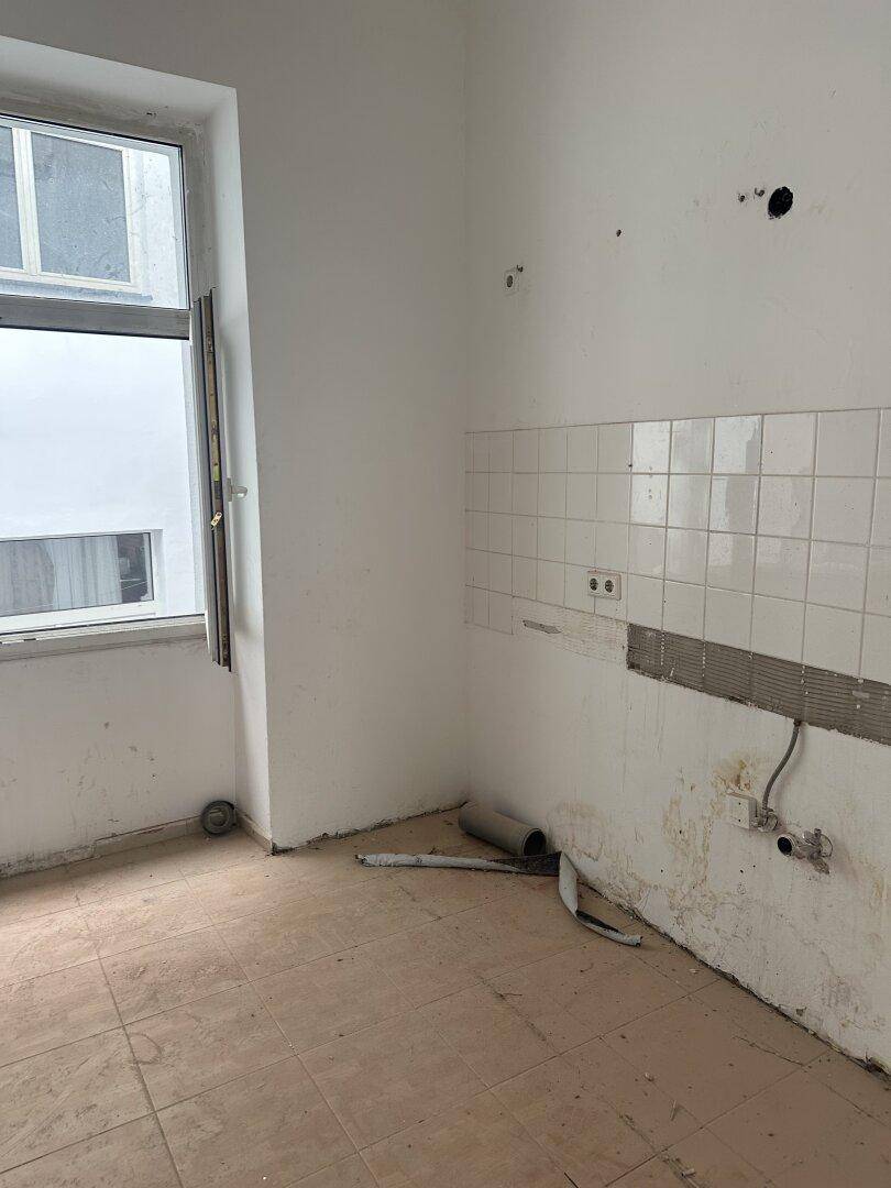 Leerer Küchenraum mit Fliesenrückwand, sichtbaren Anschlüssen und Renovierungsbedarf.