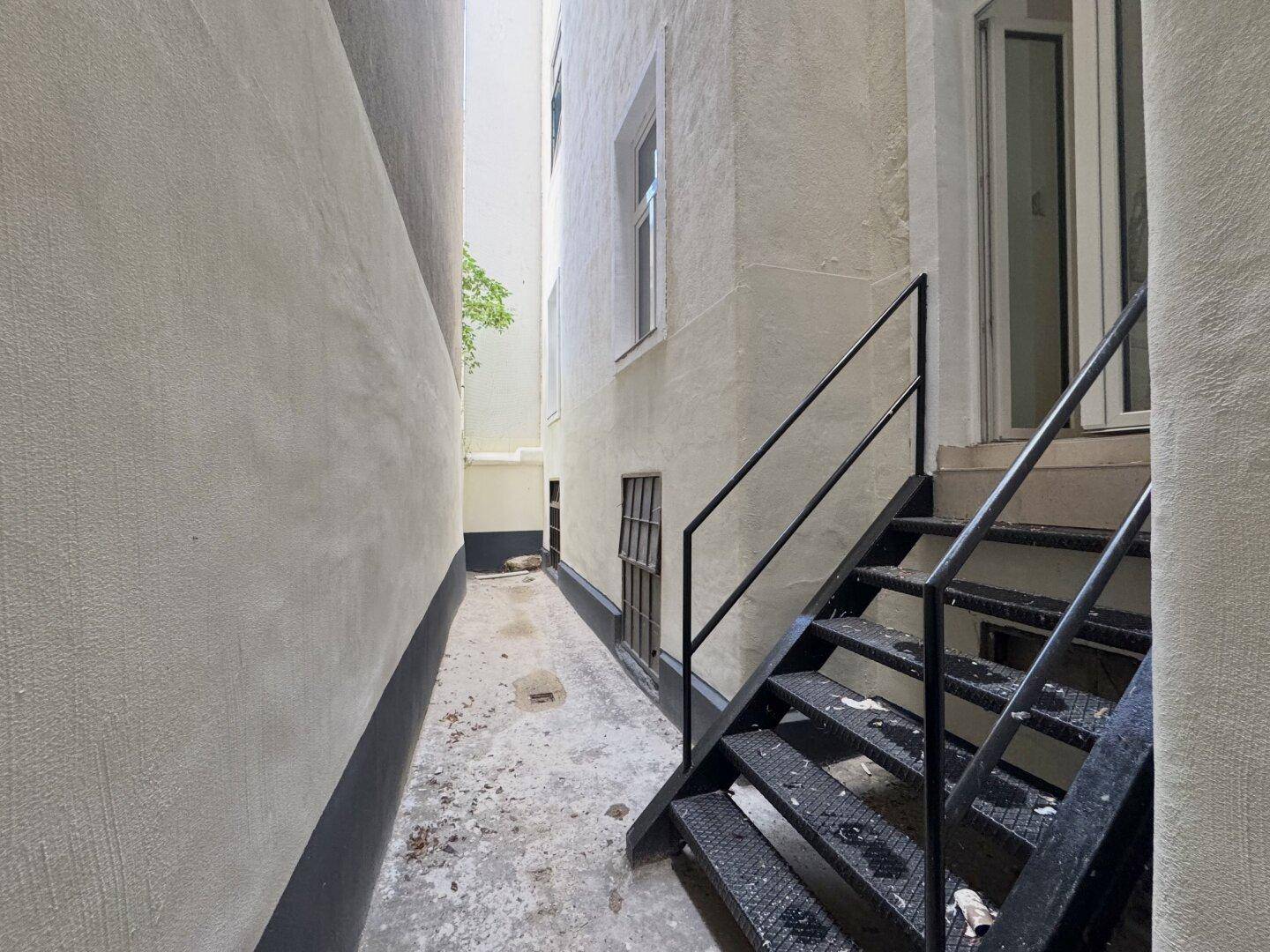 Schmaler Durchgang zwischen Gebäuden mit Außentreppe und einfacher Fassade.