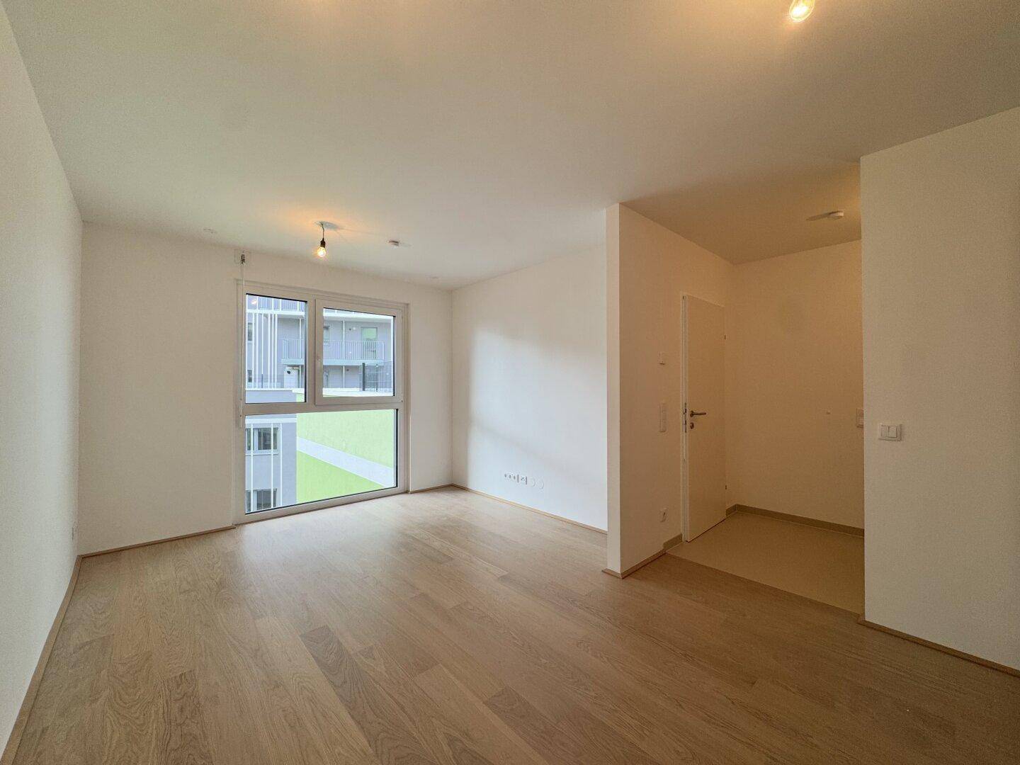 Heller Wohnraum mit großen Fenstern, Holzboden und Blick auf die Außenanlage des Gebäudes.