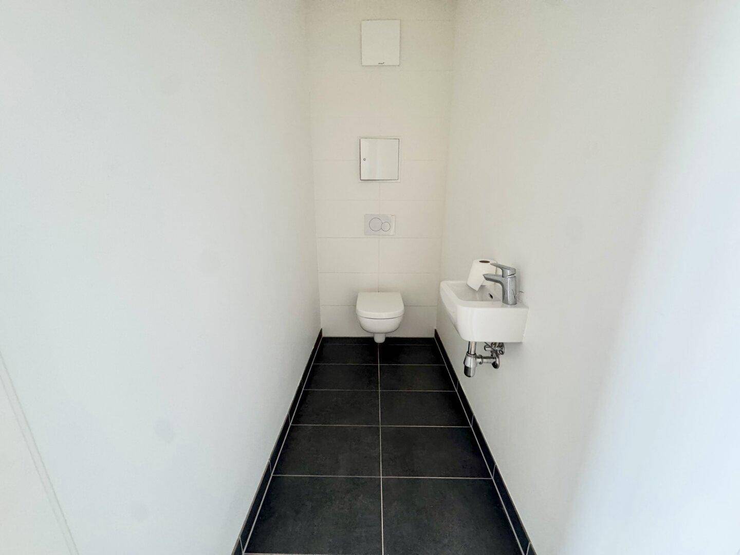 Separates WC mit wandhängender Toilette, kleinem Waschbecken und dunklen Bodenfliesen.