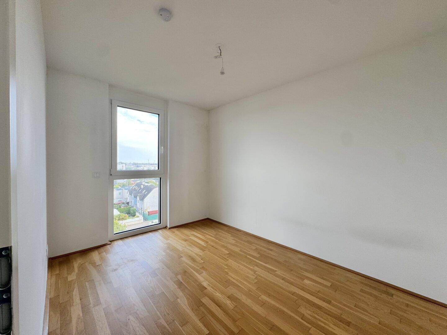 Leeres Schlafzimmer mit Parkettboden, weißen Wänden und einem großen Fenster für viel Tageslicht.