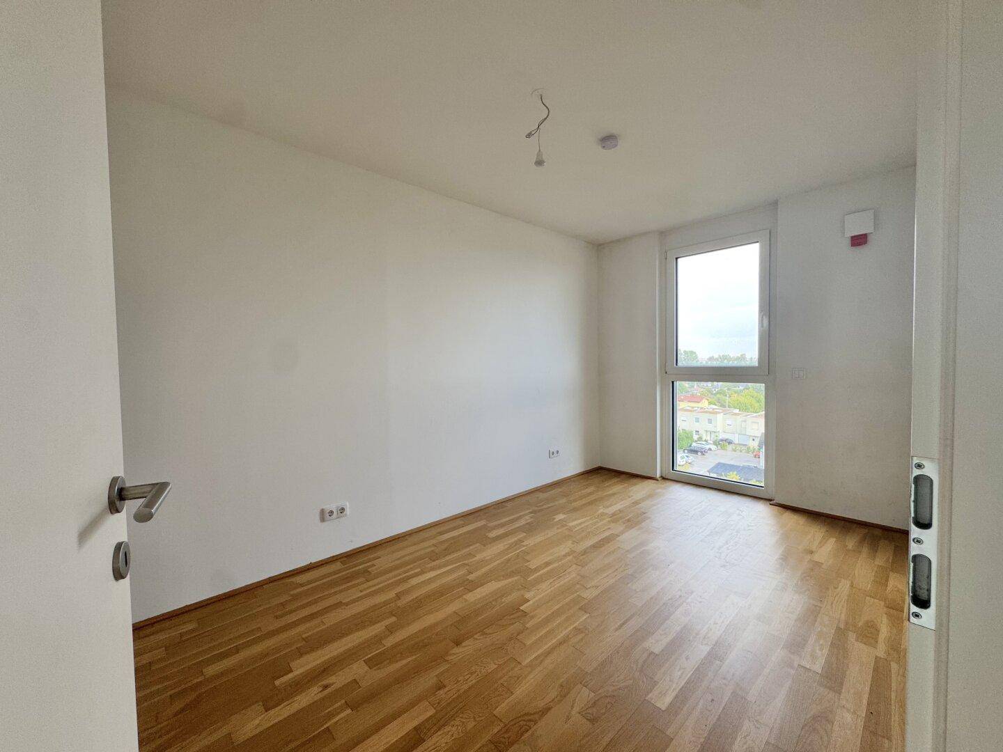 Geräumiges Schlafzimmer mit Parkettboden, weißen Wänden und einem Fenster mit Blick nach draußen.