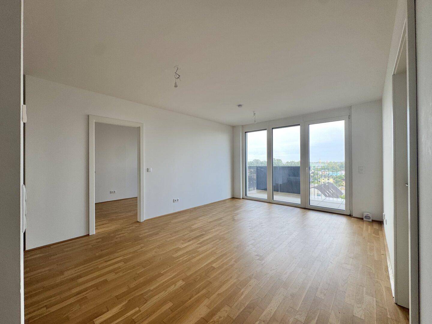 Helles Wohnzimmer mit Parkettboden, weißen Wänden und großen Fenstern zum Balkon.