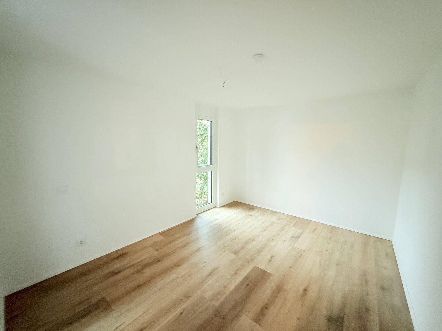 Helles Schlafzimmer mit Laminatboden und Fenster mit Blick ins Grüne.