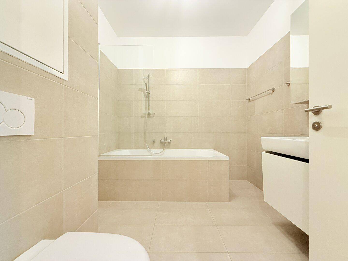 Modernes Badezimmer mit beigefarbenen Fliesen, Badewanne mit Duschfunktion und WC.