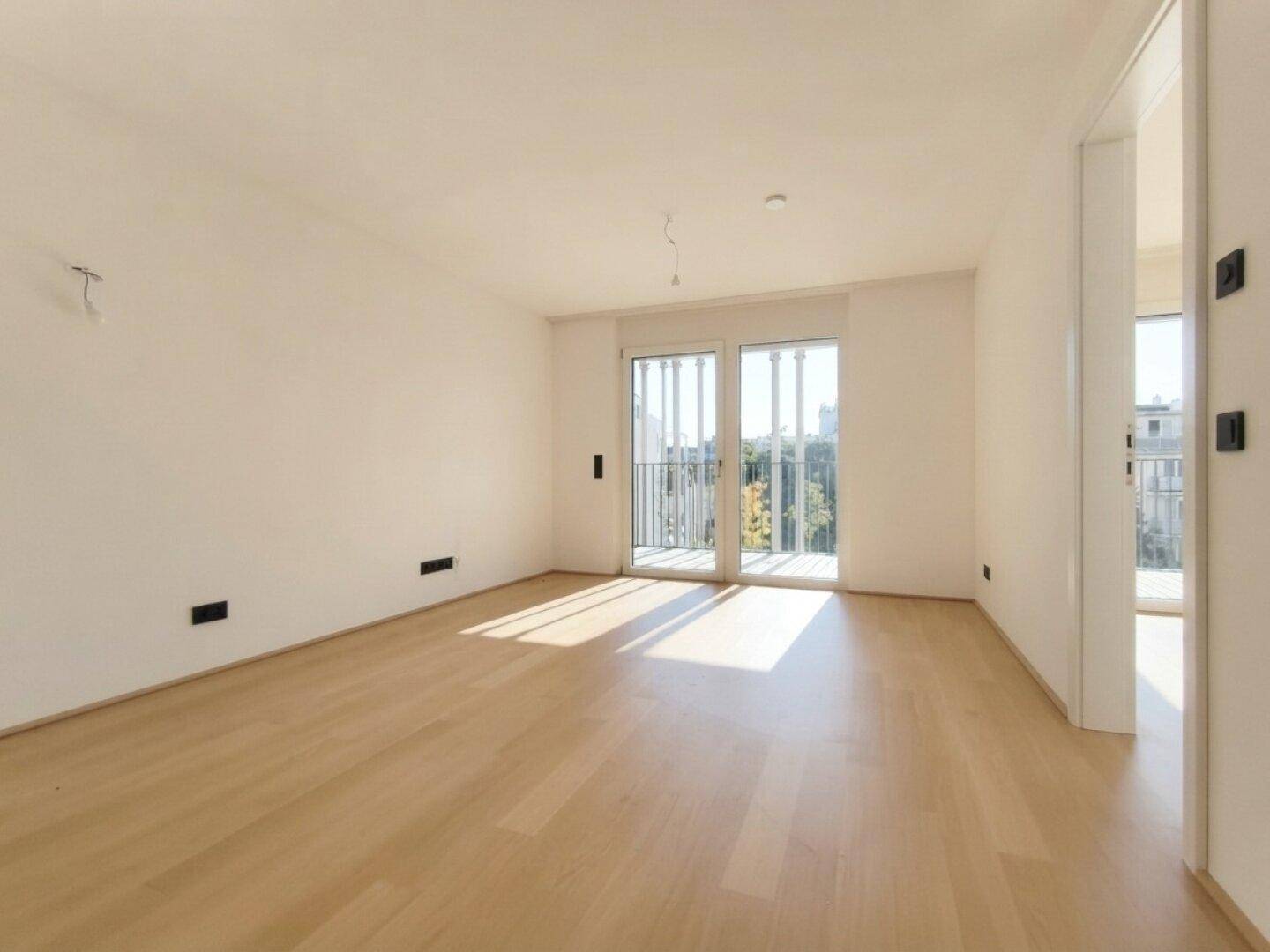 Lichtdurchfluteter Raum mit hellem Parkettboden und großen Fenstern zum Balkon.