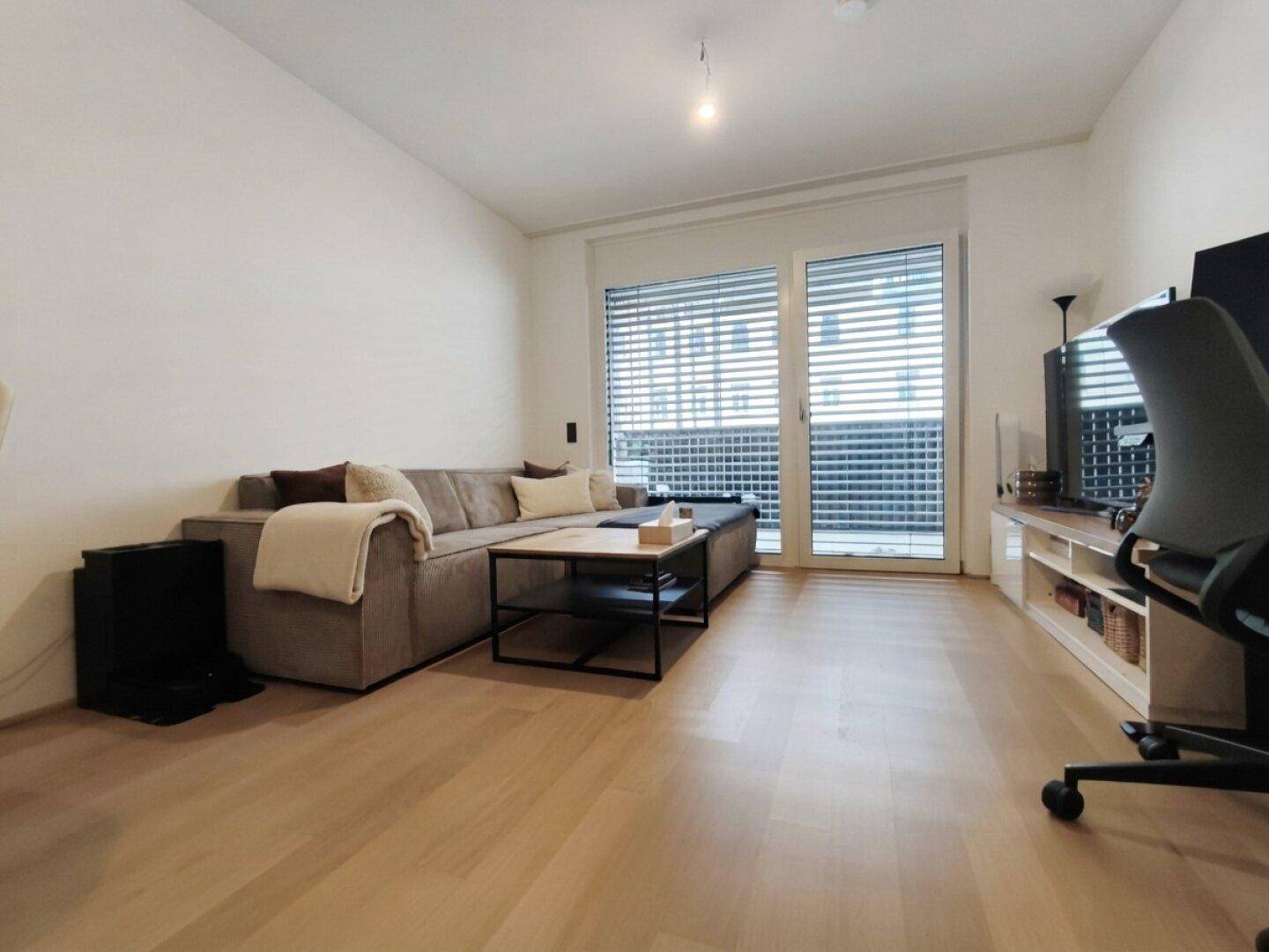 Gemütliches Wohnzimmer mit beigem Sofa, Couchtisch und großem Fenster mit Zugang zum Außenbereich.
