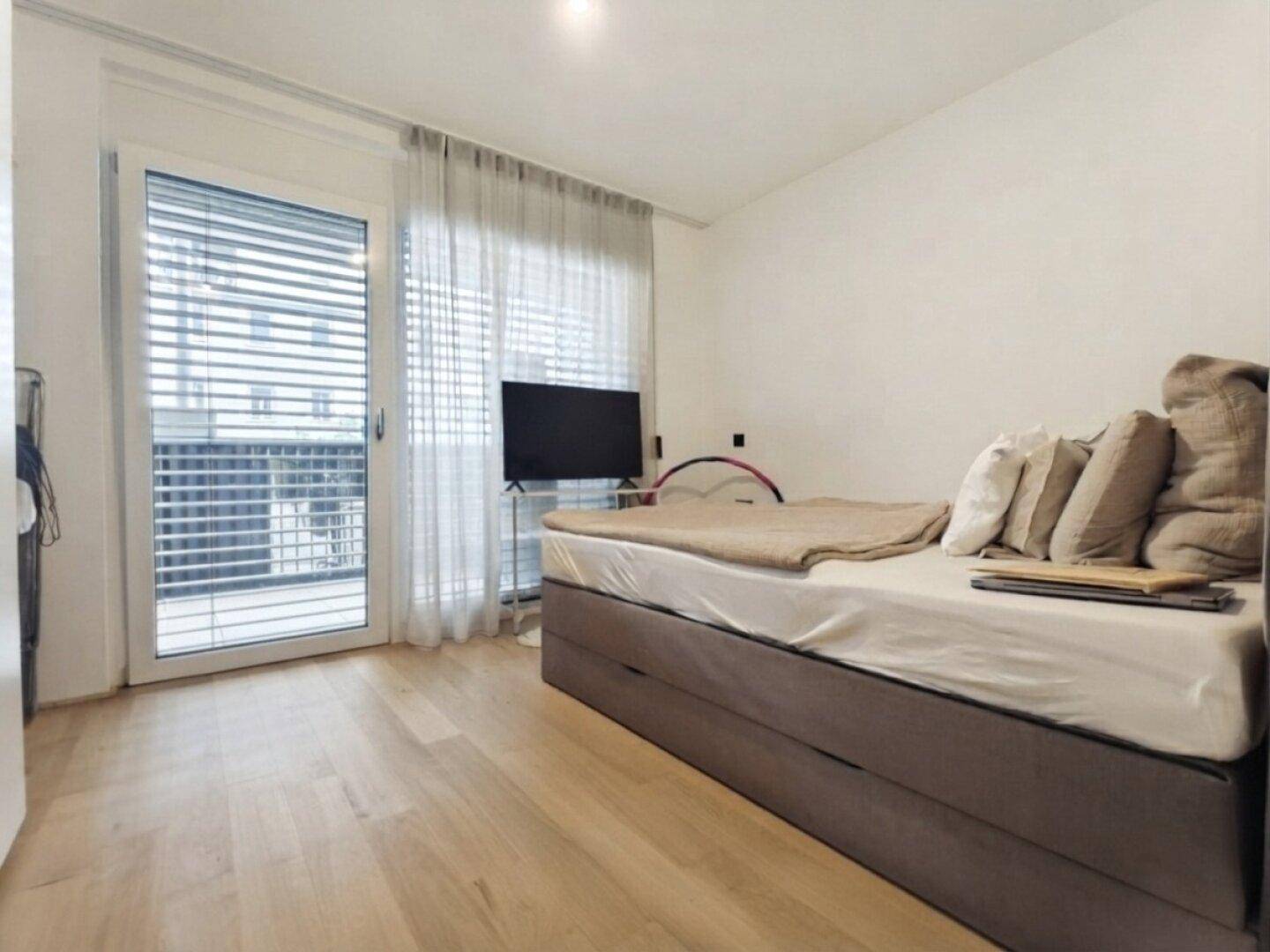 Schlafzimmer mit Bett, Fernseher und großem Fenster, das viel Tageslicht hereinlässt.