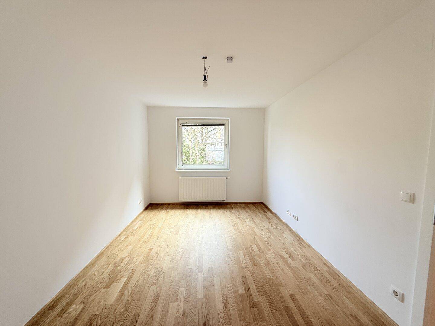 Helles, leeres Zimmer mit Holzparkettboden und einem Fenster, das viel Tageslicht hereinlässt.
