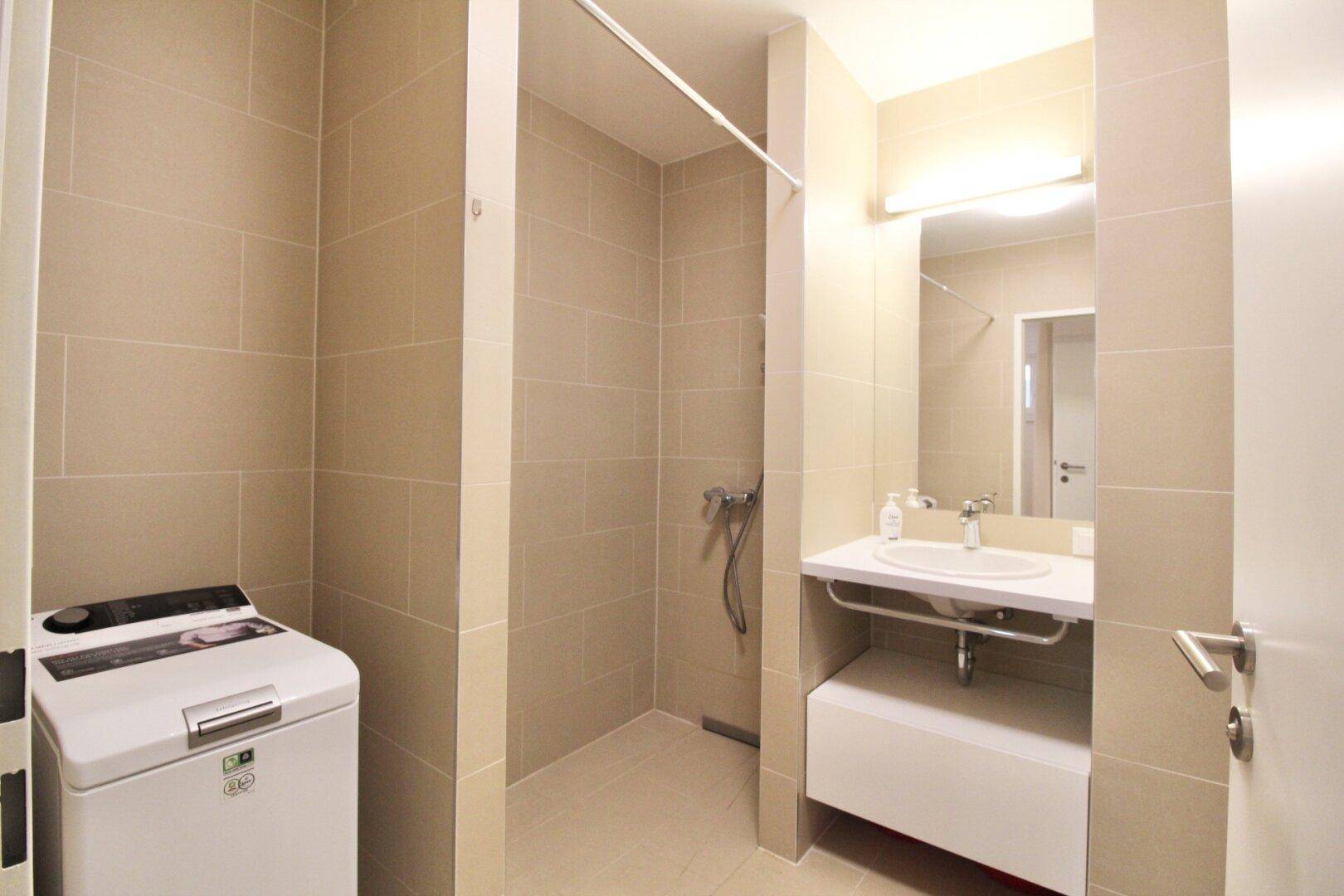 Modernes Badezimmer mit begehbarer Dusche, Waschbecken und einer Waschmaschine, hell gefliest.
