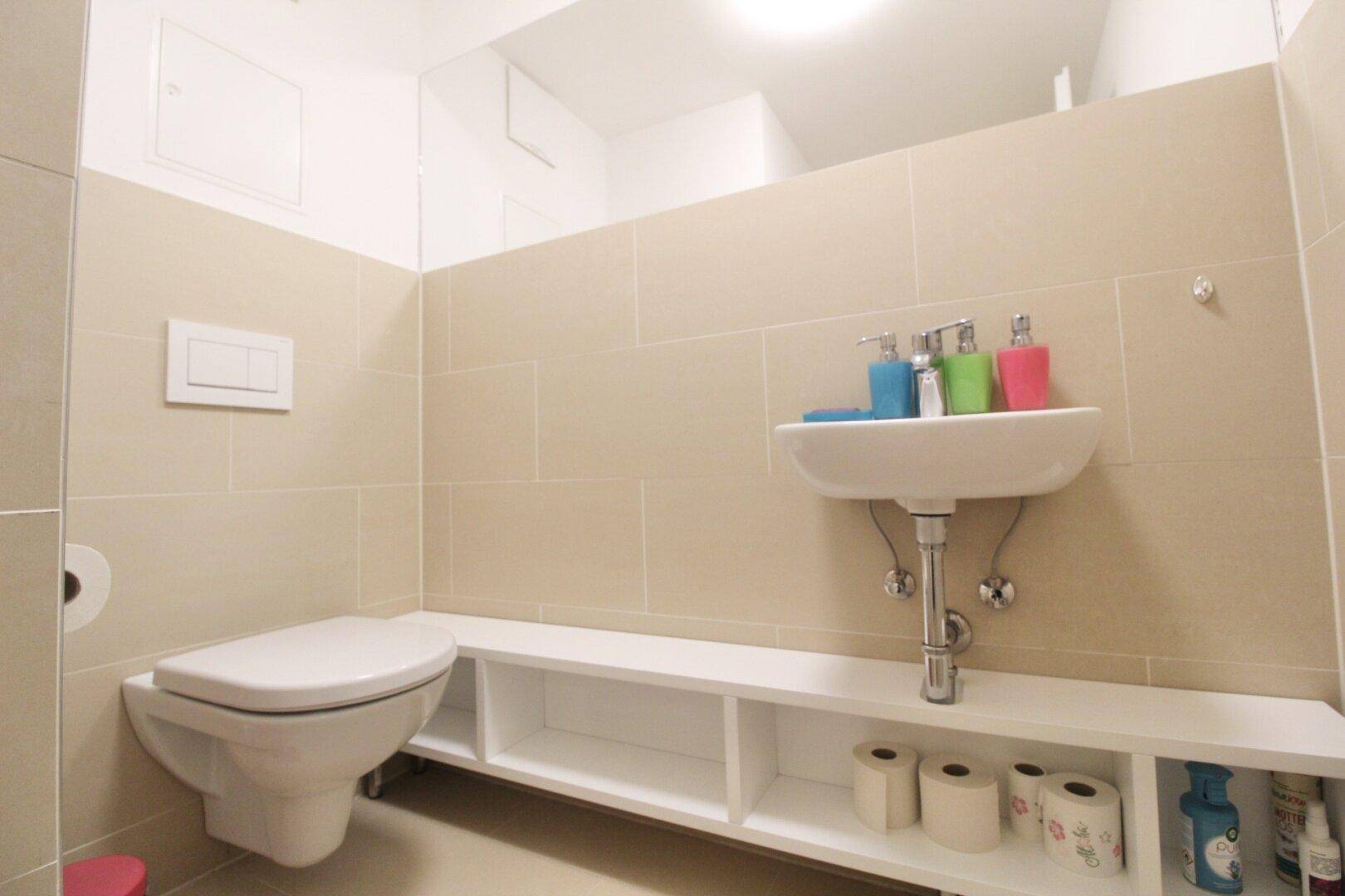 Separate Toilette mit Wand-WC, Waschbecken und integrierten Ablageflächen, modern gefliest.