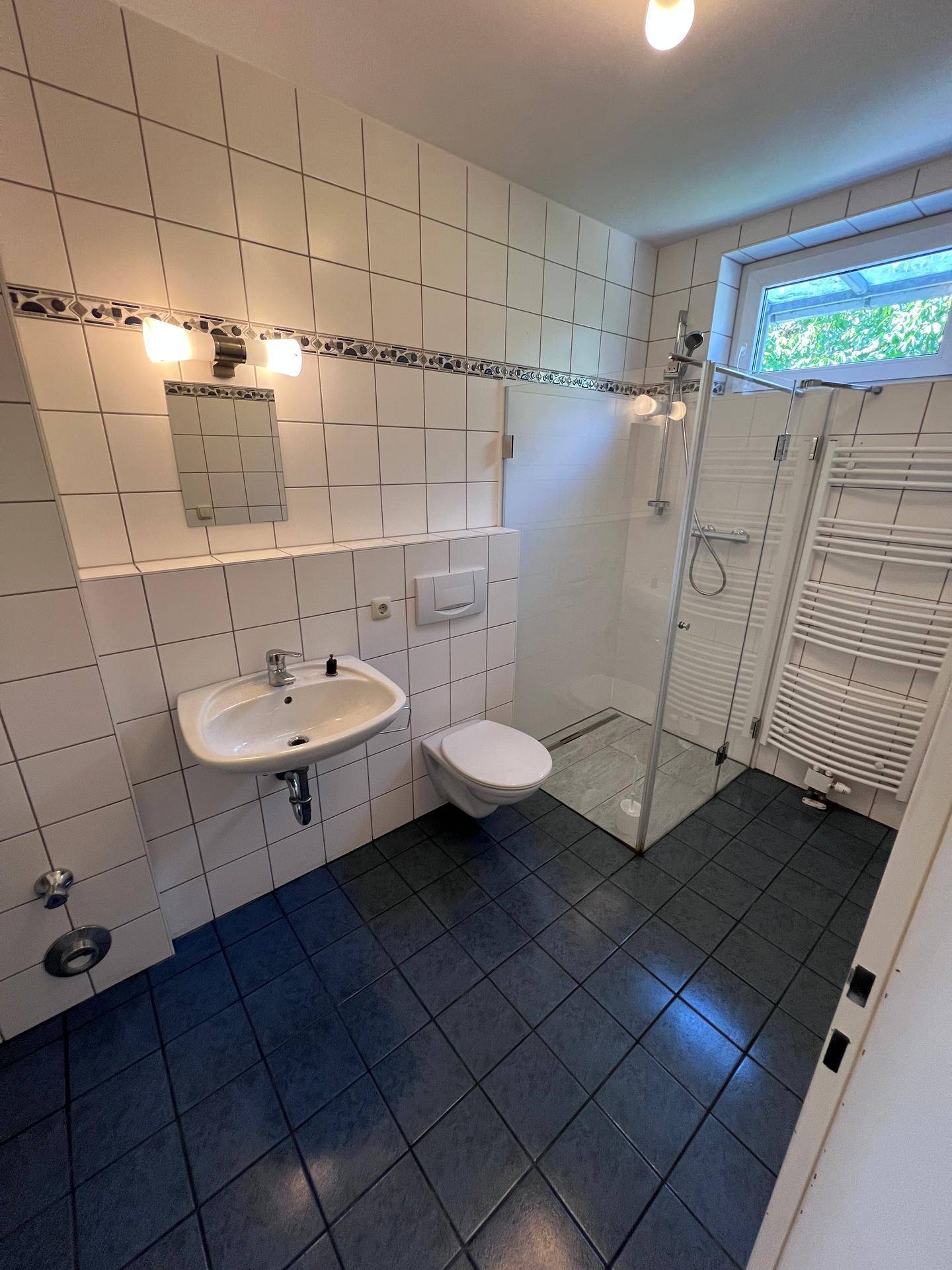 Modernes Badezimmer mit weißen Wandfliesen, dunklen Bodenfliesen, begehbarer Dusche und Fenster.