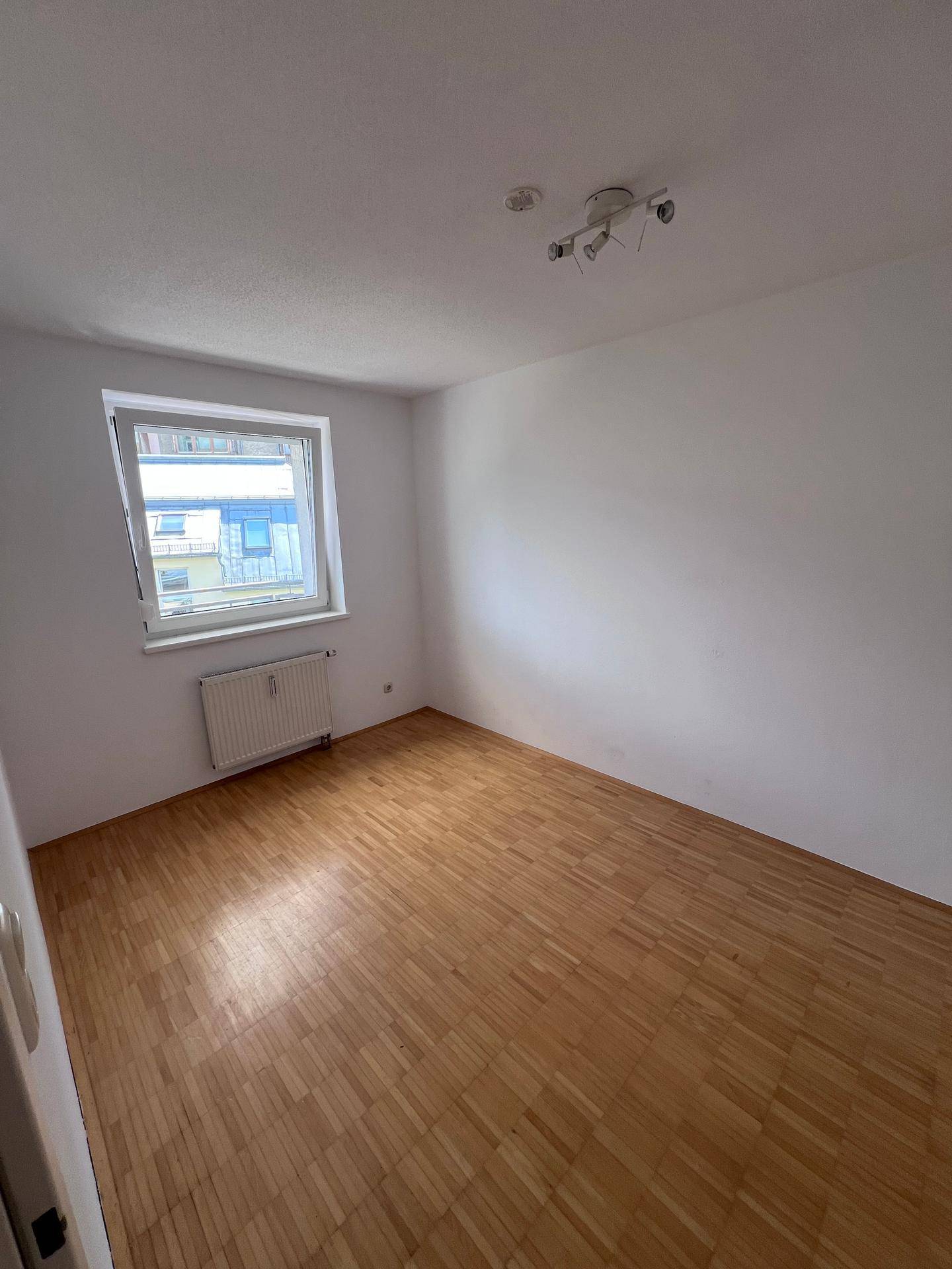 Helles, leeres Schlafzimmer mit Holzparkettboden und einem Fenster, das viel Tageslicht hereinlässt.