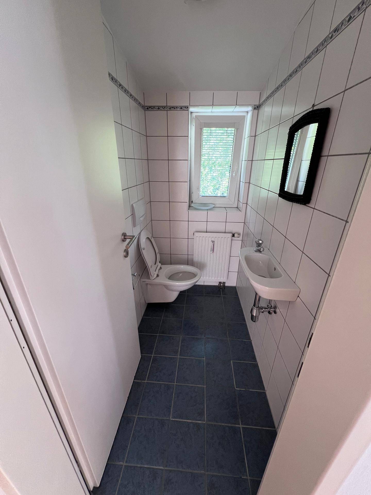 Kleines Gäste-WC mit weißen Fliesen, dunklem Boden, Toilette, Waschbecken und Fenster.