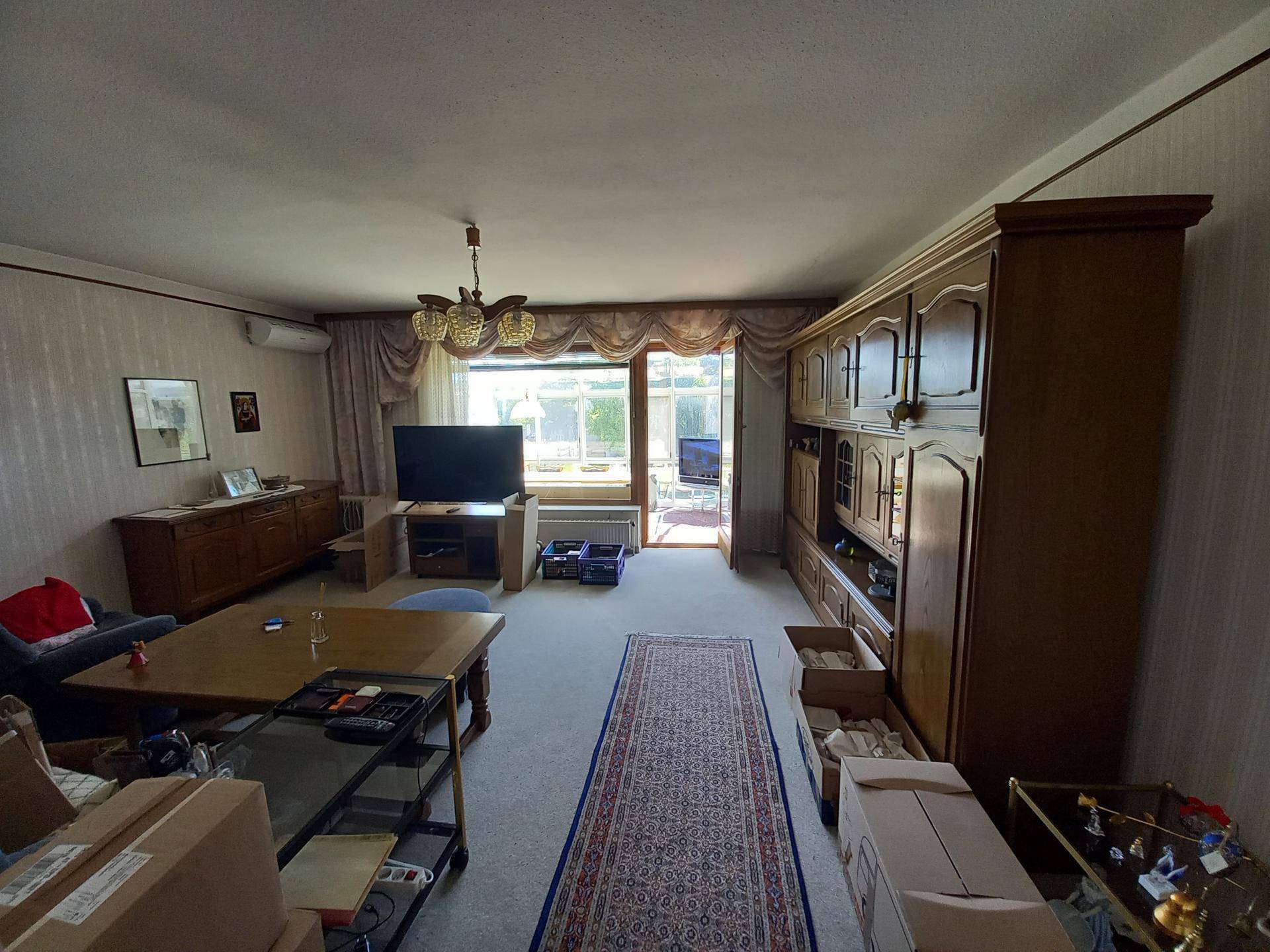 Geräumiges Wohnzimmer mit Teppichboden, traditionellen Holzmöbeln und Zugang zu einem Außenbereich.