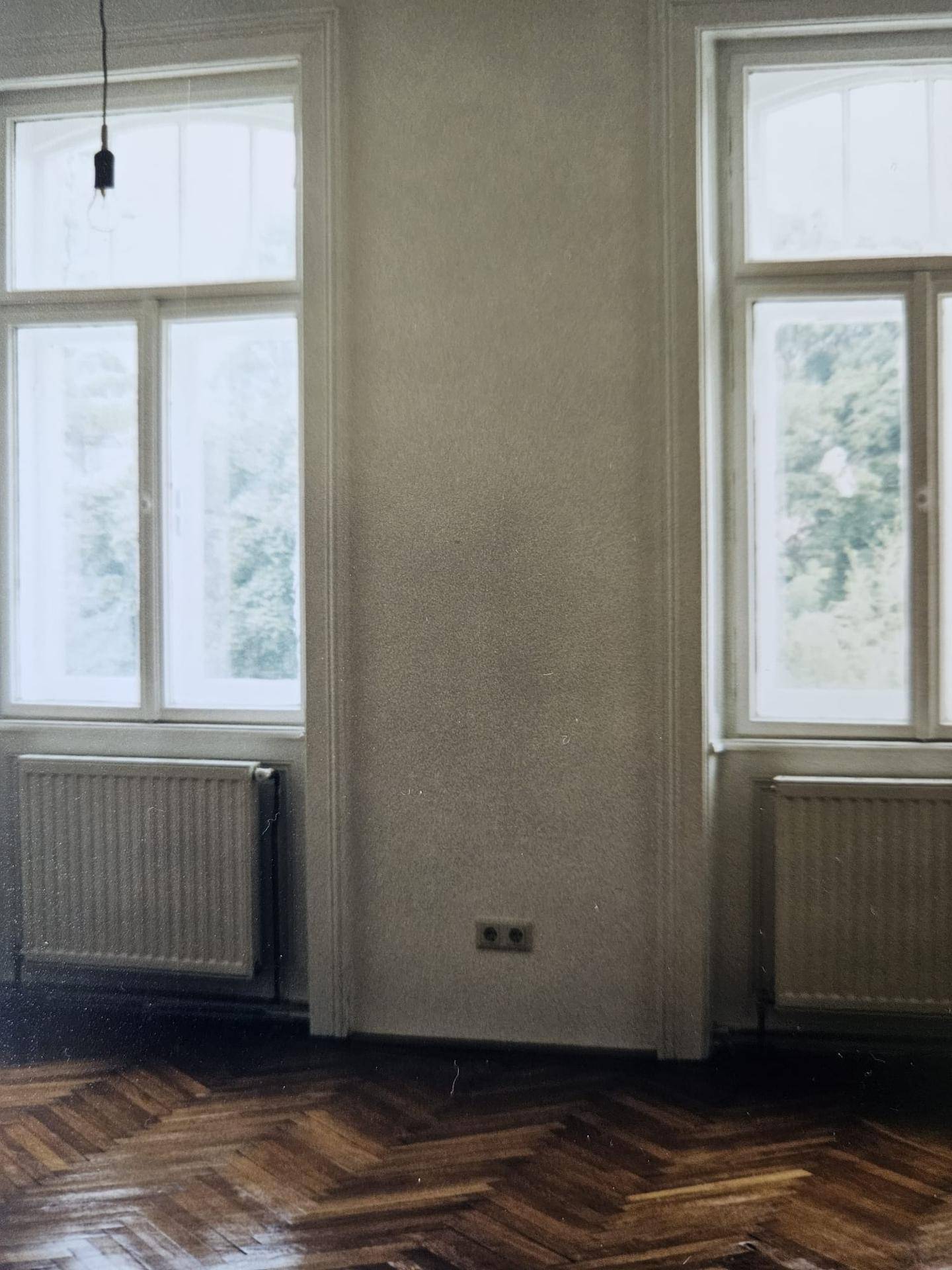 Helles Zimmer mit zwei großen Fenstern, Heizkörpern und dunklem Fischgrätparkettboden.
