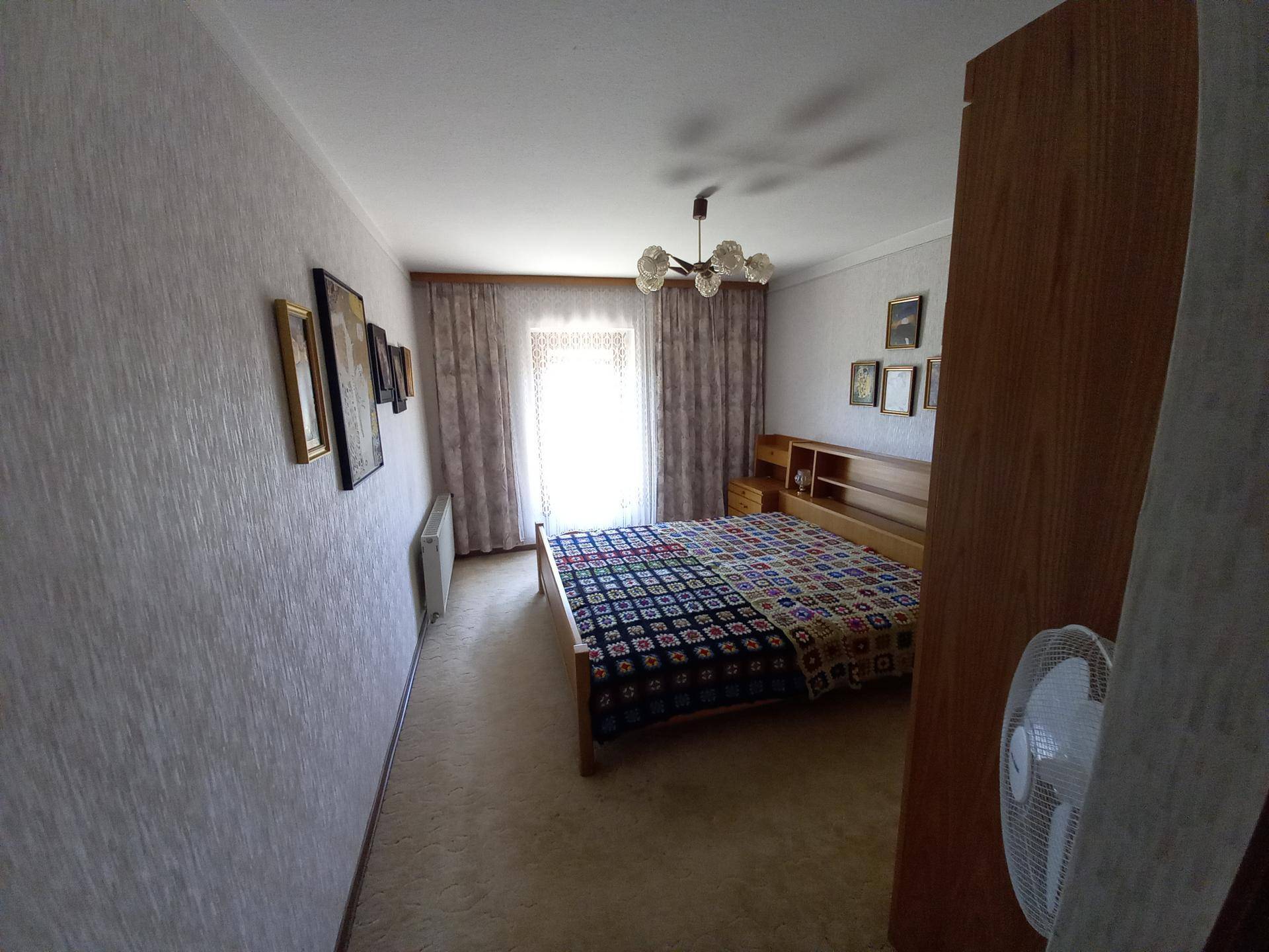 Schlafzimmer mit Doppelbett, Teppichboden und Fenster mit Vorhängen, das viel Tageslicht hereinlässt.