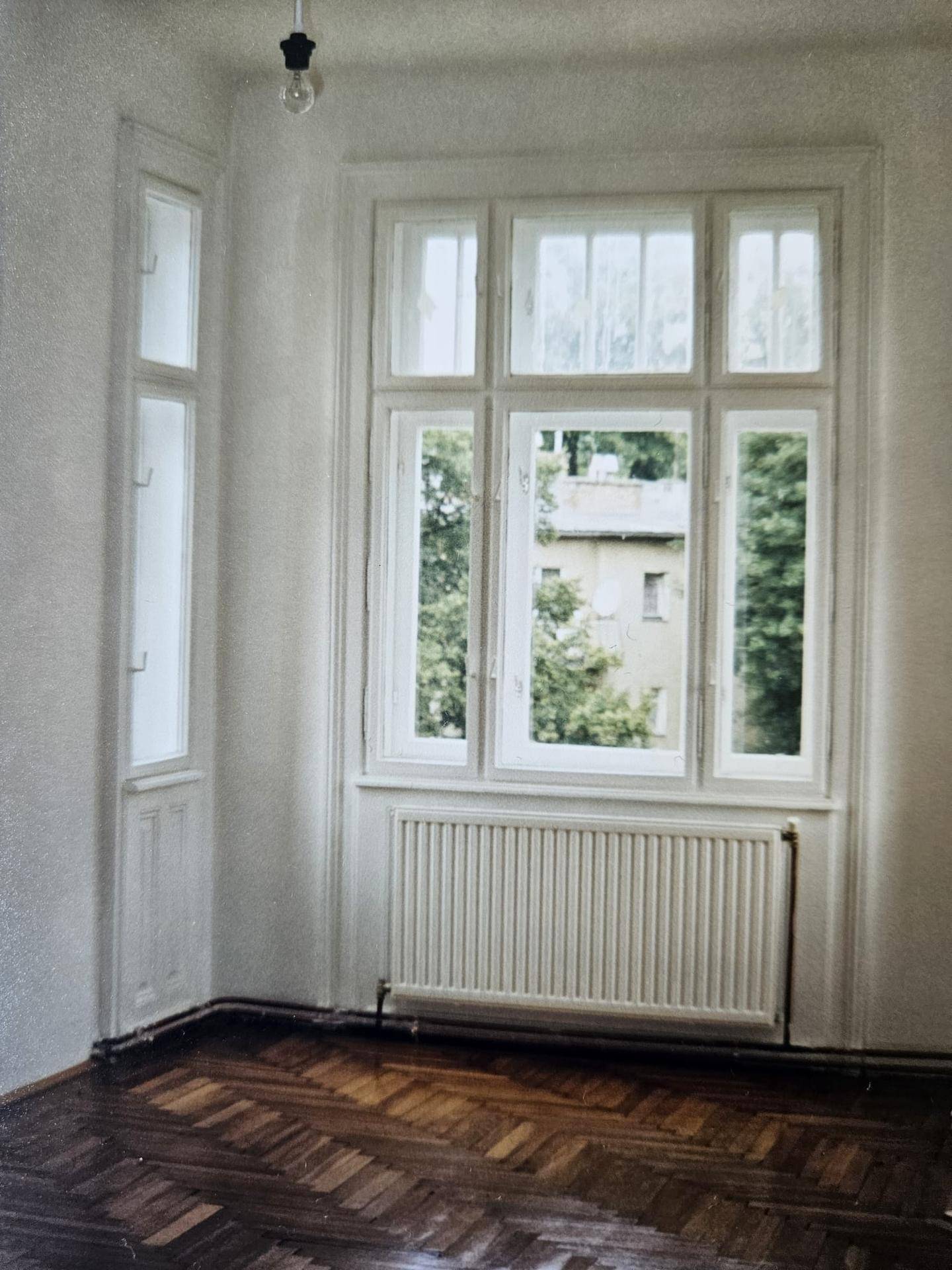 Geräumiges Zimmer mit dunklem Fischgrätparkett, großem Fenster und Heizkörper, Blick ins Grüne.