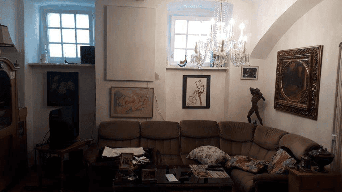 Wohnzimmer mit einem großen Ecksofa, zwei Fenstern und einem opulenten Kronleuchter unter einer Gewölbedecke.