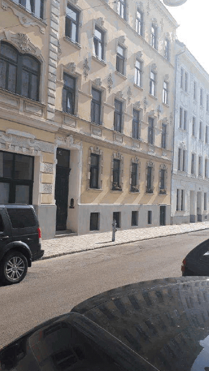 Historisches Gebäude mit reich verzierten Fassadendetails und parkenden Autos auf der Straße an einem sonnigen Tag.