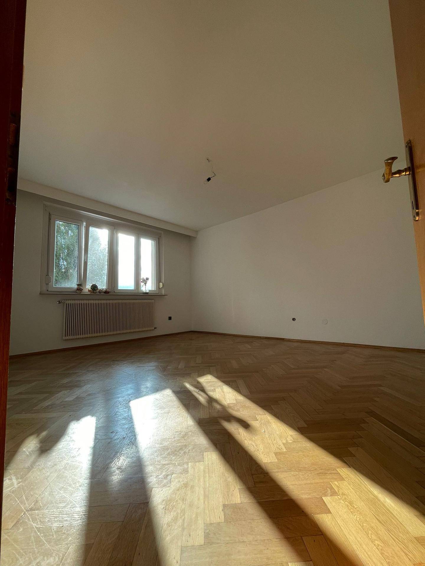 Geräumiges Zimmer mit Parkettboden, Fenster und Heizkörper, durchflutet von Sonnenlicht.
