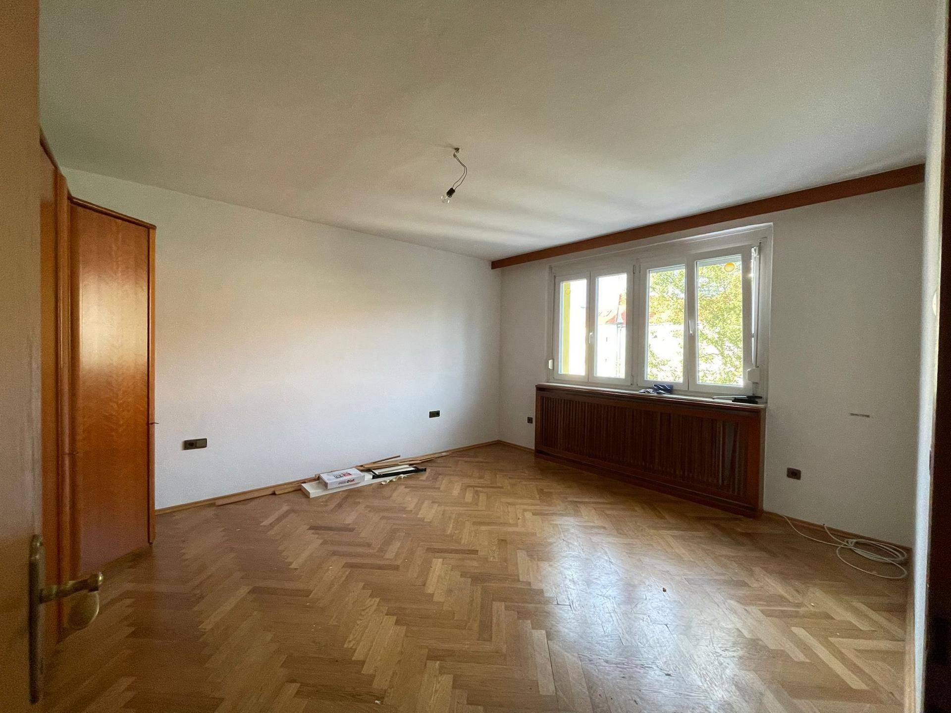 Geräumiges Schlafzimmer mit Parkettboden, großem Fenster und Holzverkleidung am Heizkörper.