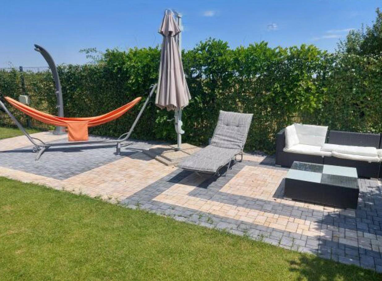 Gemütliche gepflasterte Terrasse mit Gartenmöbeln, Hängematte und Sonnenschirm, umgeben von einer grünen Hecke.