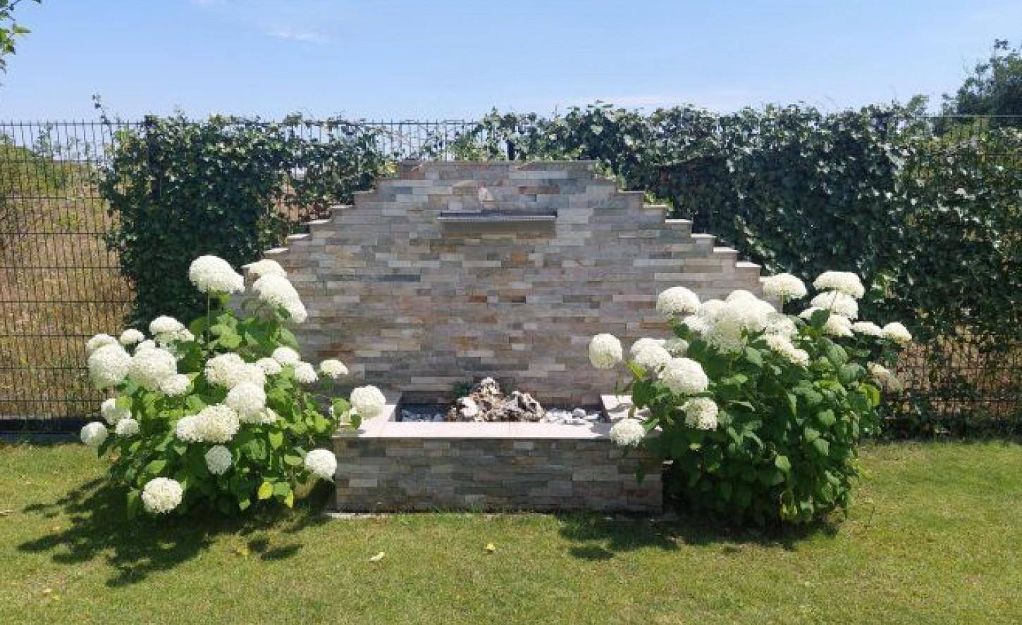 Dekorativer Steinbrunnen im Garten, umgeben von blühenden Hortensien und gepflegtem Rasen.