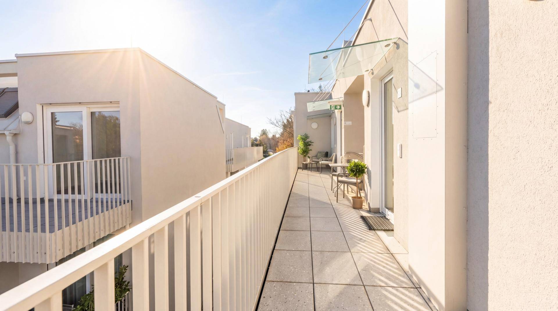 Langer, sonniger Balkon mit Fliesenboden und Blick auf benachbarte Gebäude.
