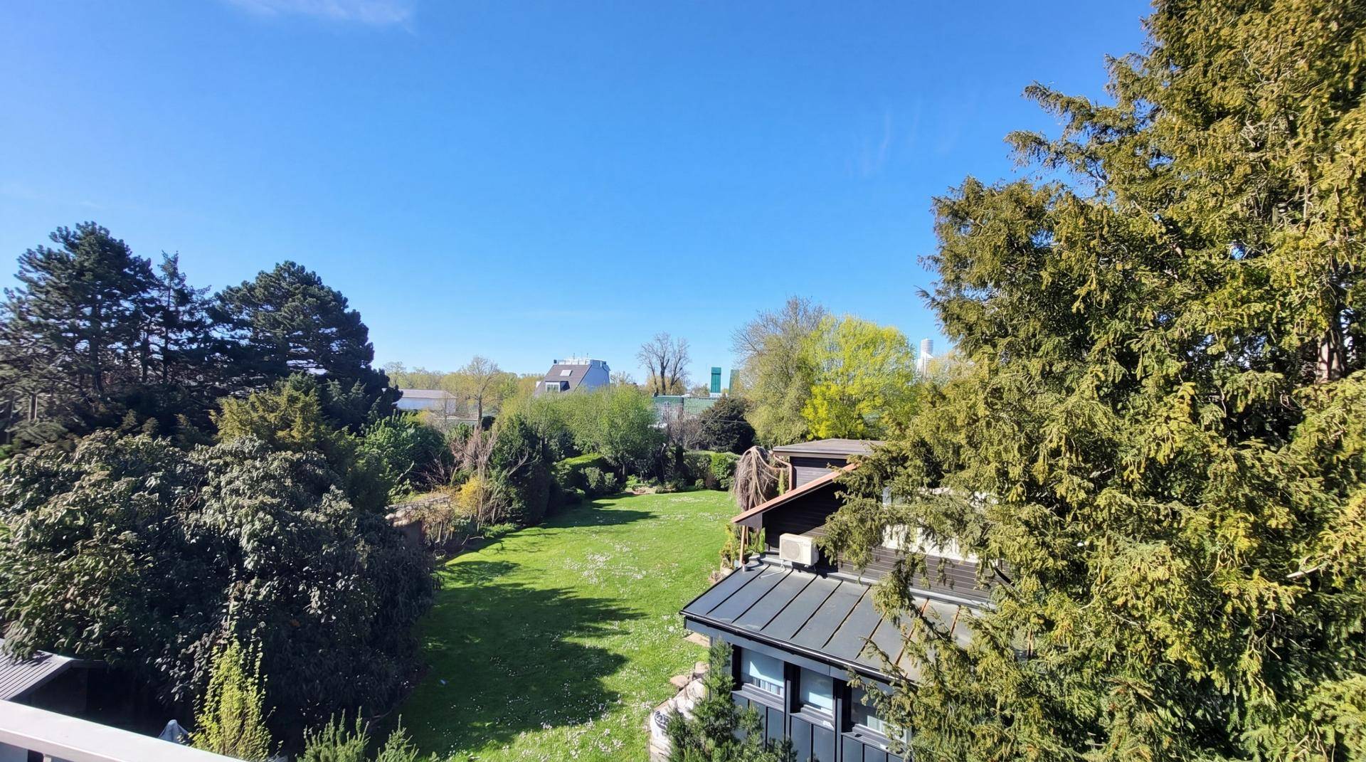 Weitläufiger Blick über grüne Gärten und benachbarte Häuser unter blauem Himmel.