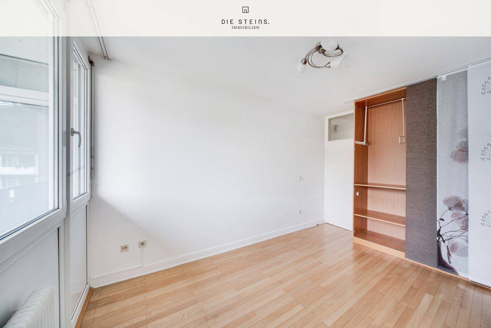 Schlafzimmer mit Parkettboden, großem Fenster und integriertem Schrankbereich, ideal für Stauraum.