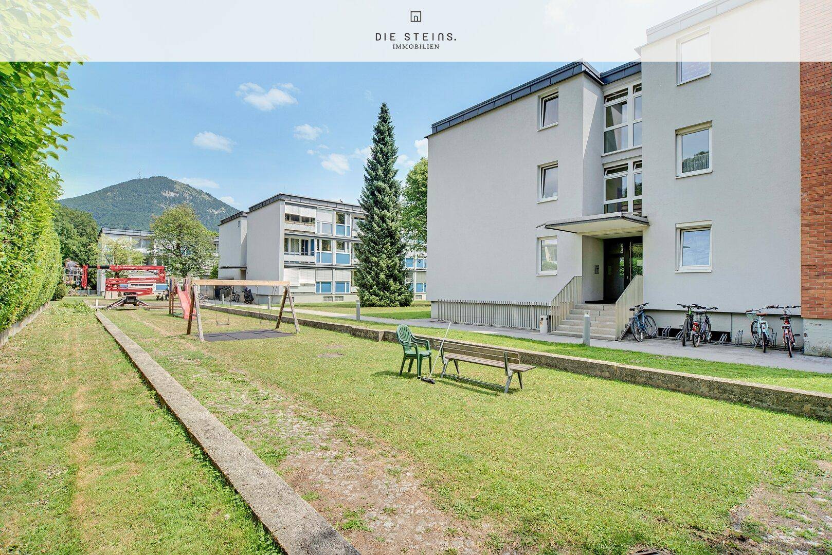 Gemeinschaftlicher Gartenbereich mit Rasenfläche, Spielplatz und Sitzgelegenheiten, umgeben von Bäumen.