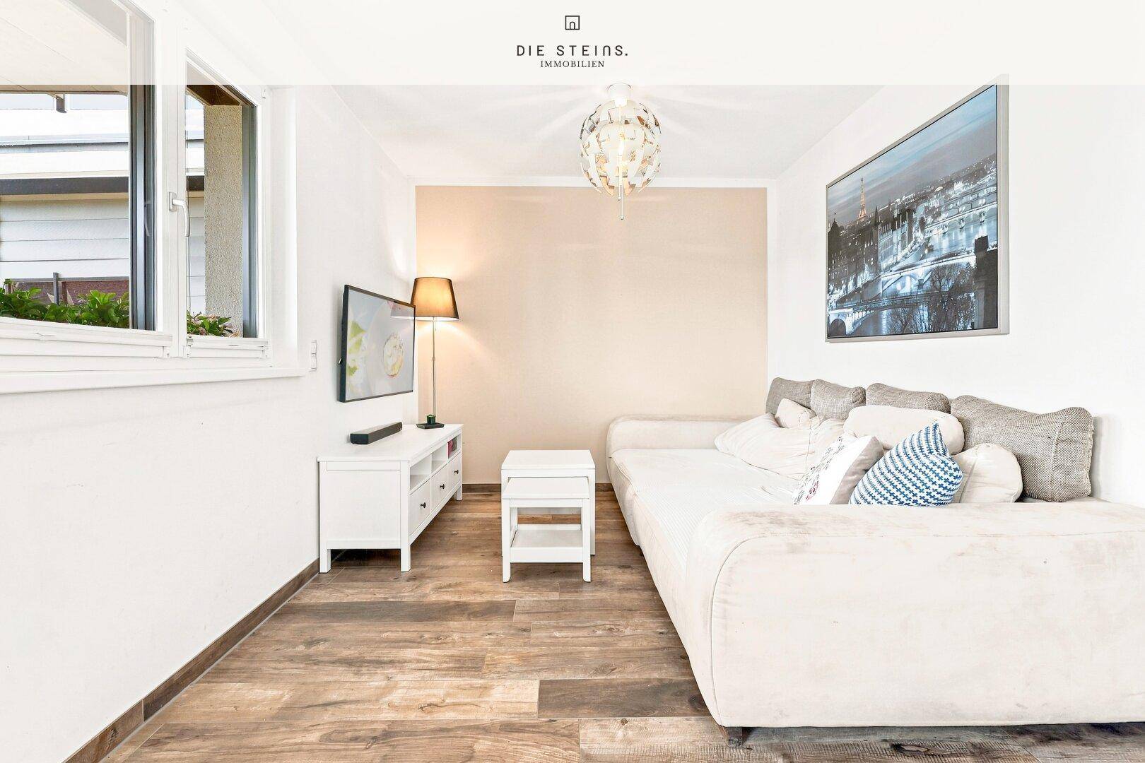 Gemütliches Wohnzimmer mit hellem Sofa, Fernseher und Holzoptik-Boden.
