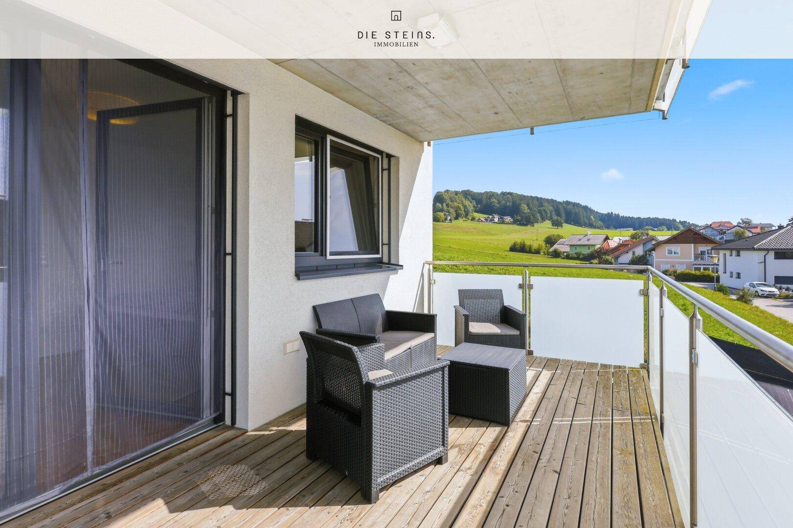 Sonniger Balkon mit Holzbelag, Sitzmöbeln und weitem Blick ins Grüne.