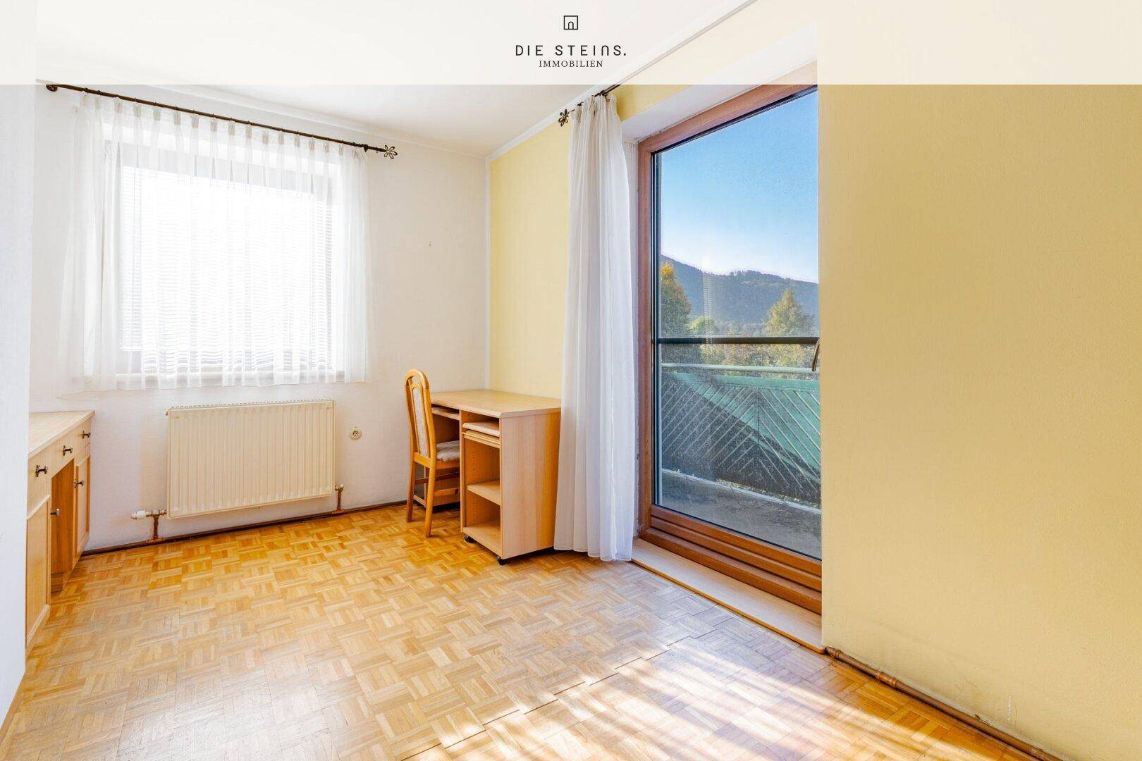 Helles Zimmer mit Parkettboden, Schreibtisch und Zugang zu einem Balkon.
