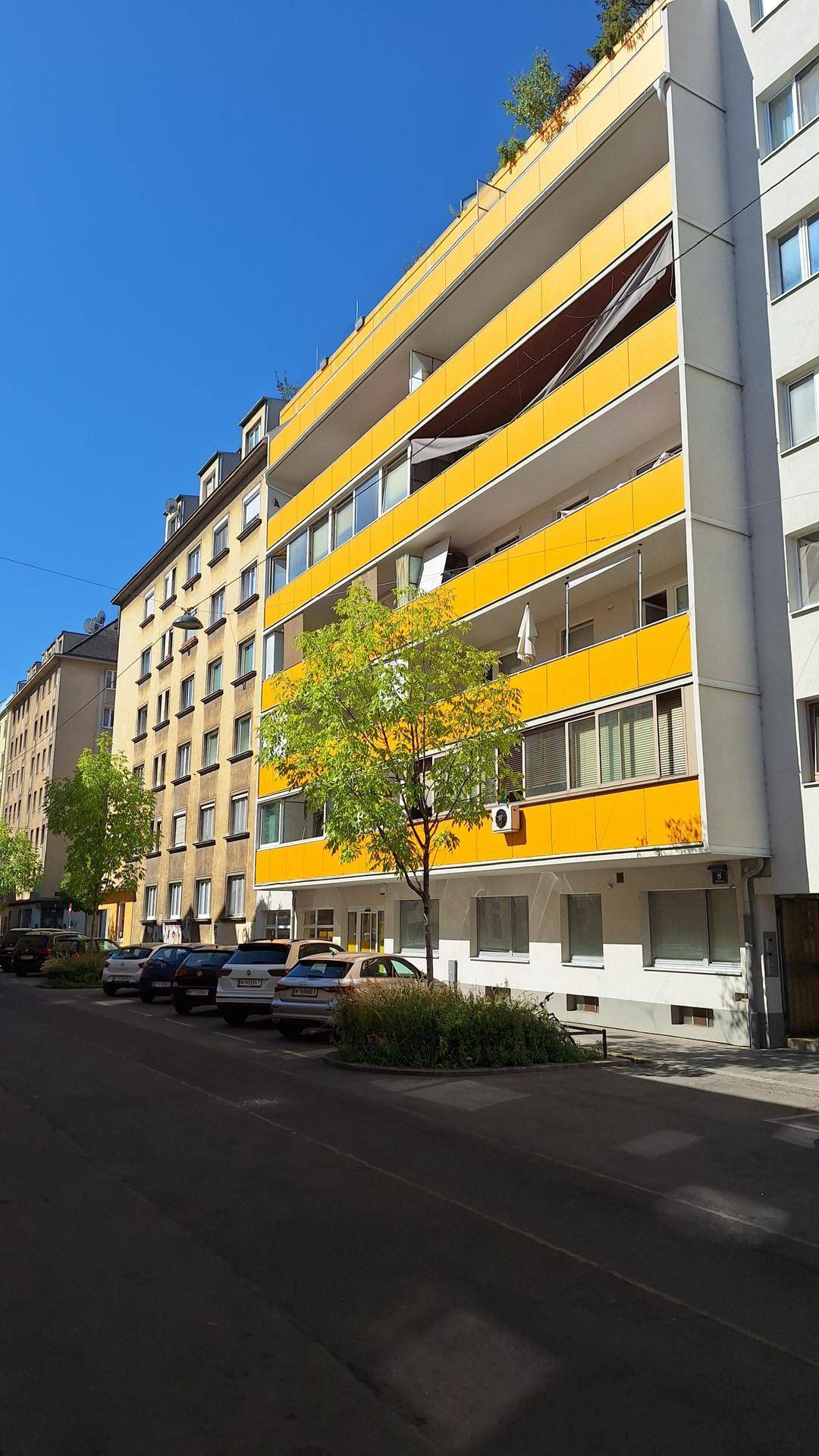 Ansicht eines modernen Gebäudes mit gelben Balkonen und umliegenden Häusern an einer sonnigen Straße.