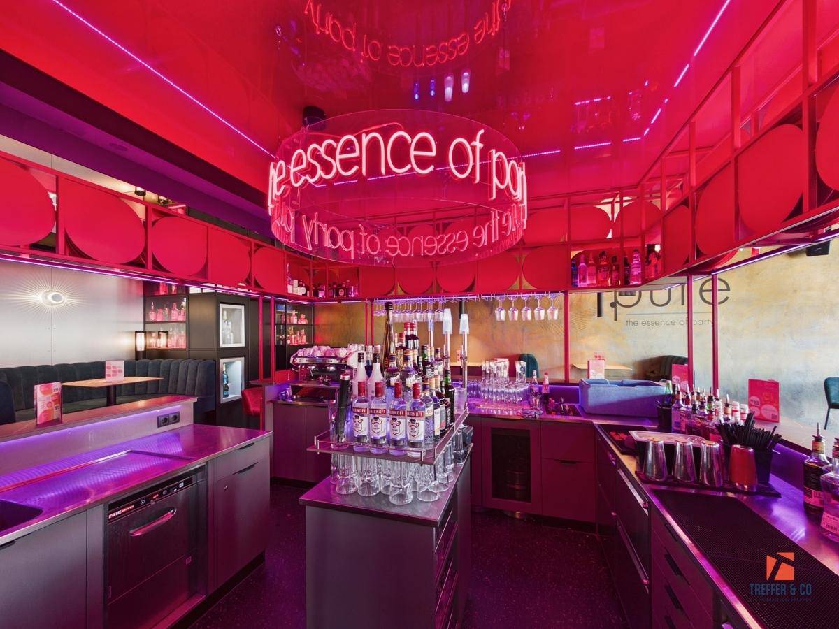 Blick hinter die Bar mit professionellen Geräten, zahlreichen Flaschen und einem leuchtenden Schriftzug.