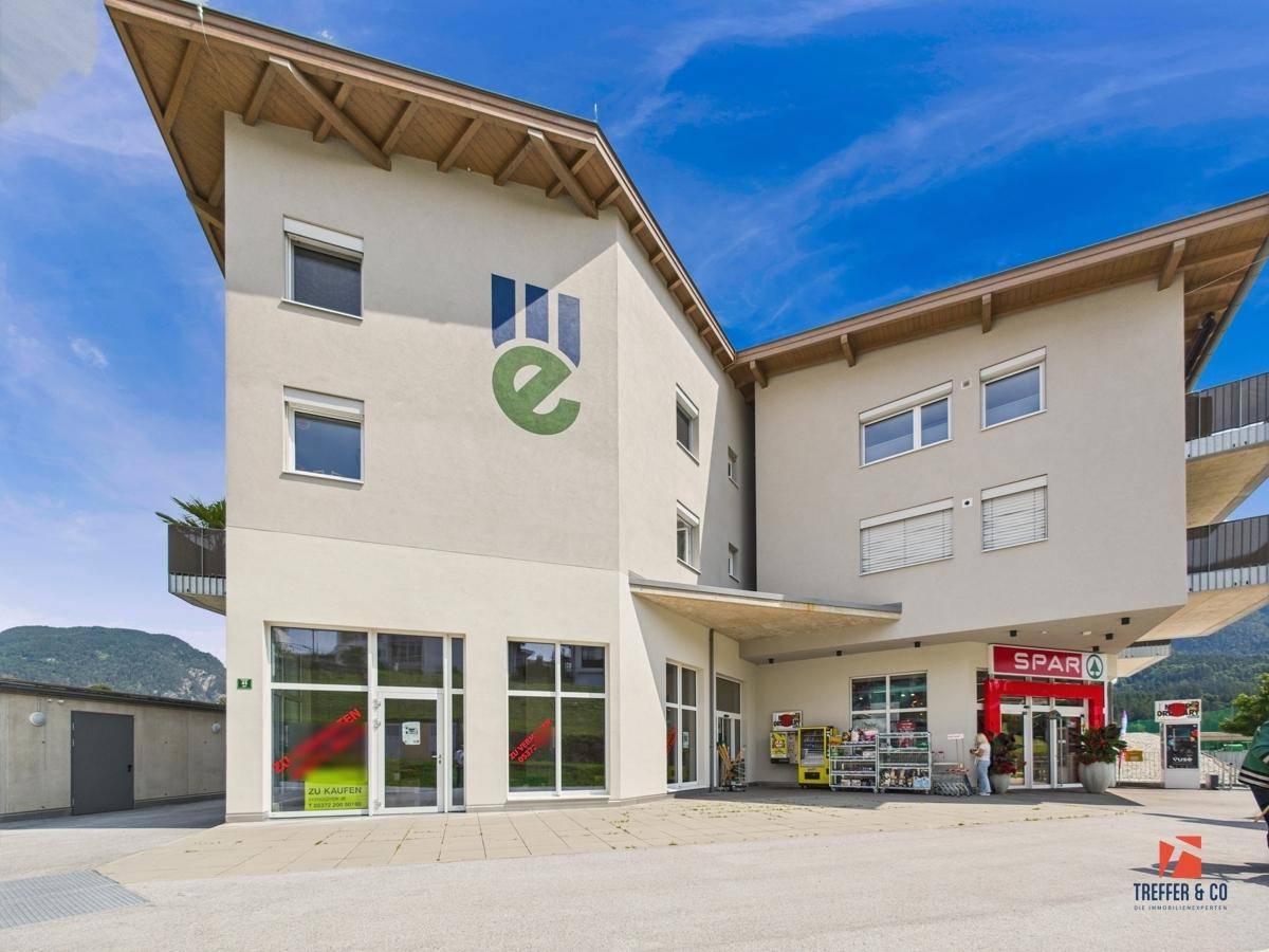 Modernes Geschäftsgebäude mit großen Schaufenstern, Supermarkt und Blick auf die umliegende Berglandschaft.
