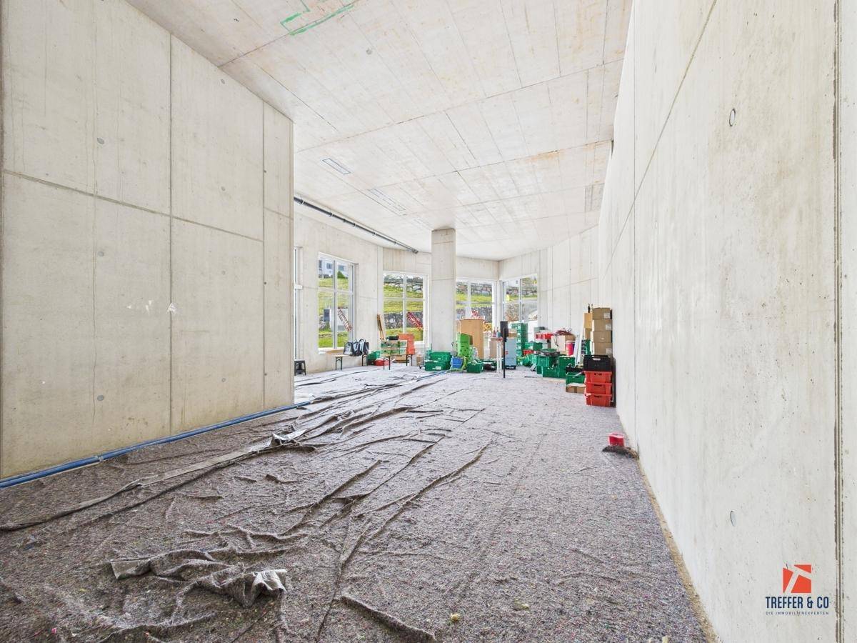 Geräumiger Gewerberaum im Rohbau mit großen Fenstern und Blick auf die grüne Umgebung.