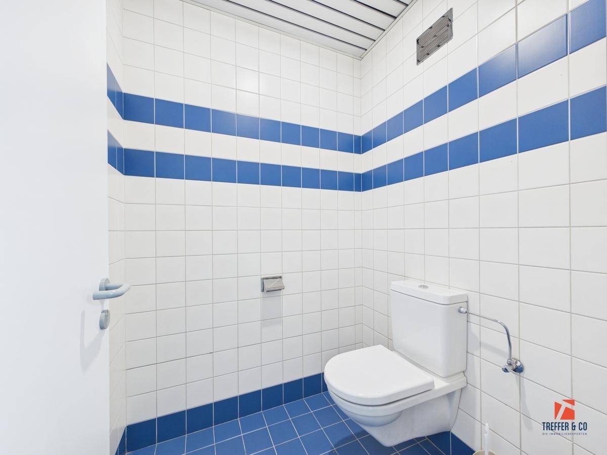 Toilettenraum mit weiß-blauen Fliesen, einer Wandtoilette und einem Lüftungsgitter an der Decke.