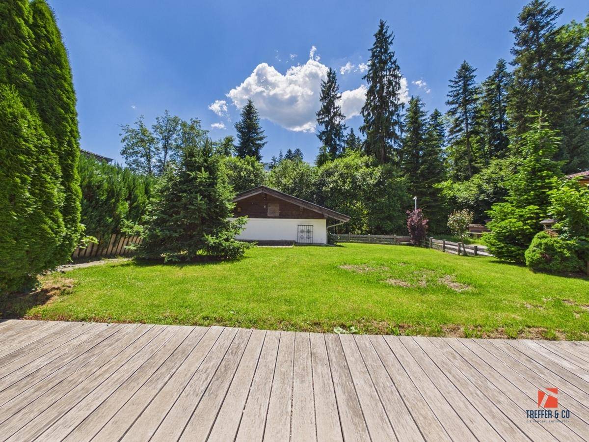 Gepflegter Garten mit Rasenfläche und Holzterrasse, umgeben von hohen Bäumen.