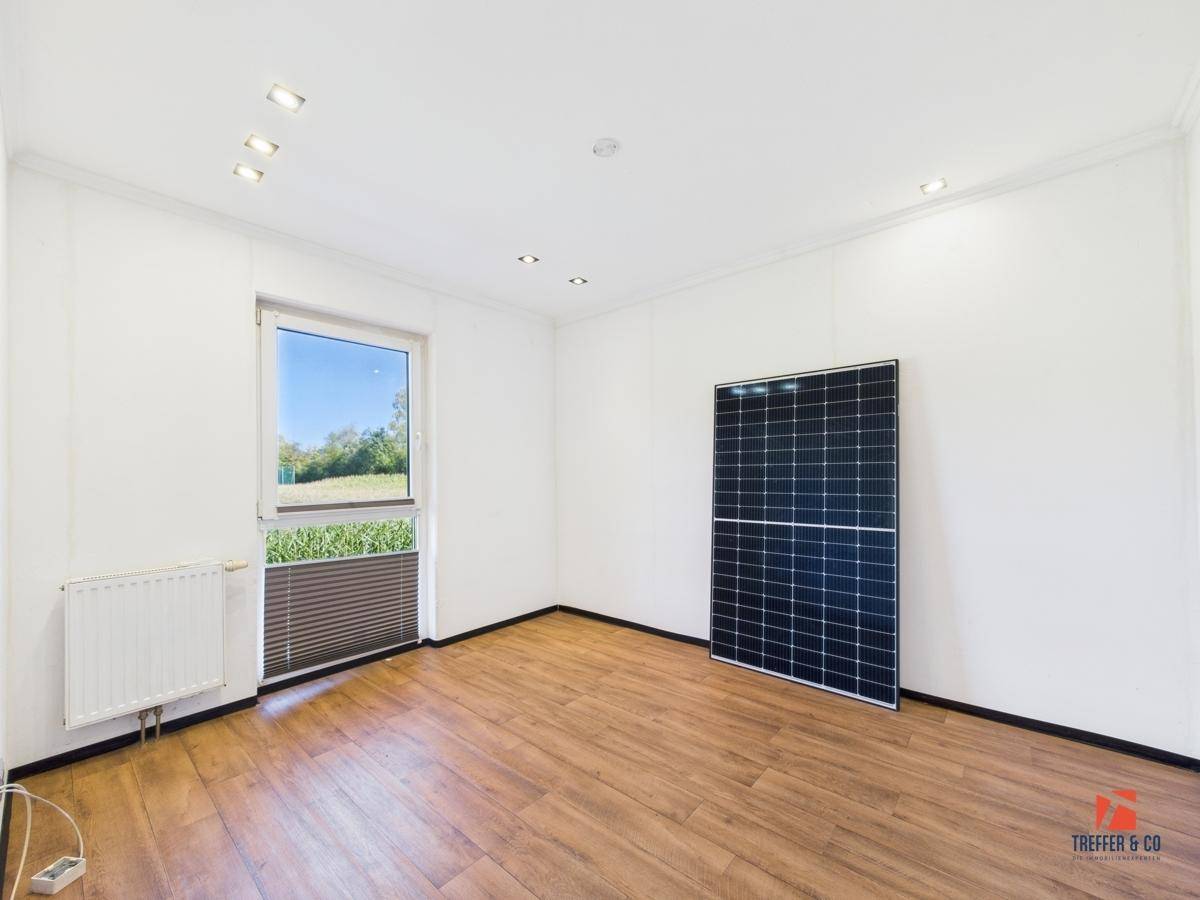 Helles Zimmer mit Laminatboden, Fenster und einem Solarpaneel an der Wand.